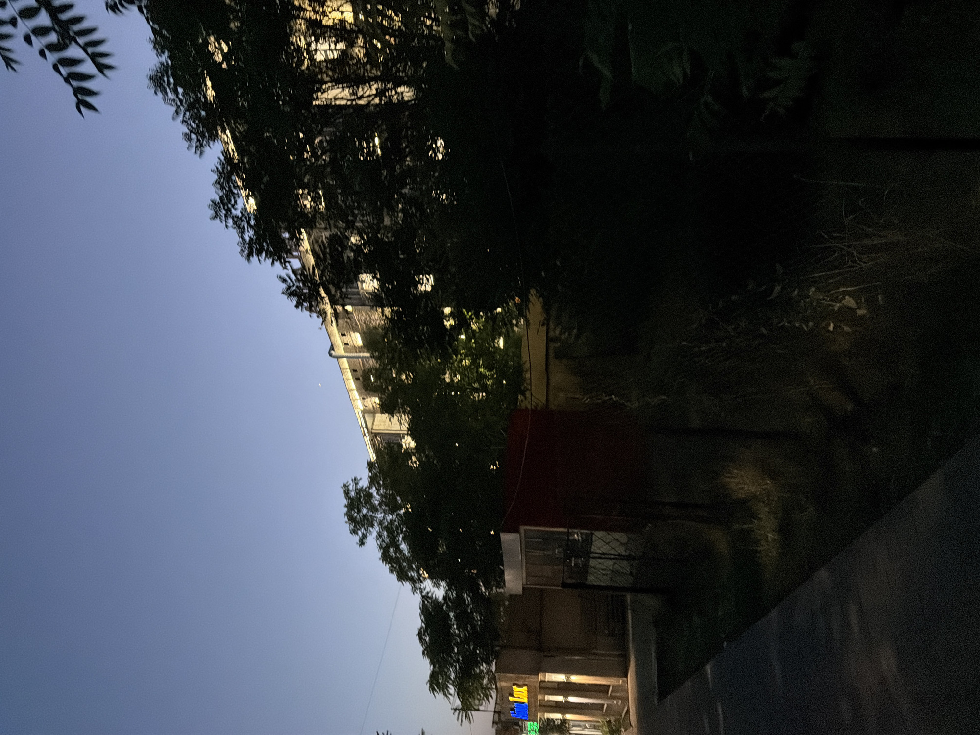 The image shows a street scene at dusk or dawn in Shymkent, Kazakhstan. A multi-story building with illuminated windows is visible in the background, partially obscured by dark trees. In the foreground, a sidewalk runs along the left side of the frame, leading towards a low-rise building with illuminated signage that reads "Alan Lux" in neon letters. Adjacent to this is a red structure and a fenced area. The sky is a deep blue, indicative of twilight. The overall impression is of an urban setting at the transition between day and night.