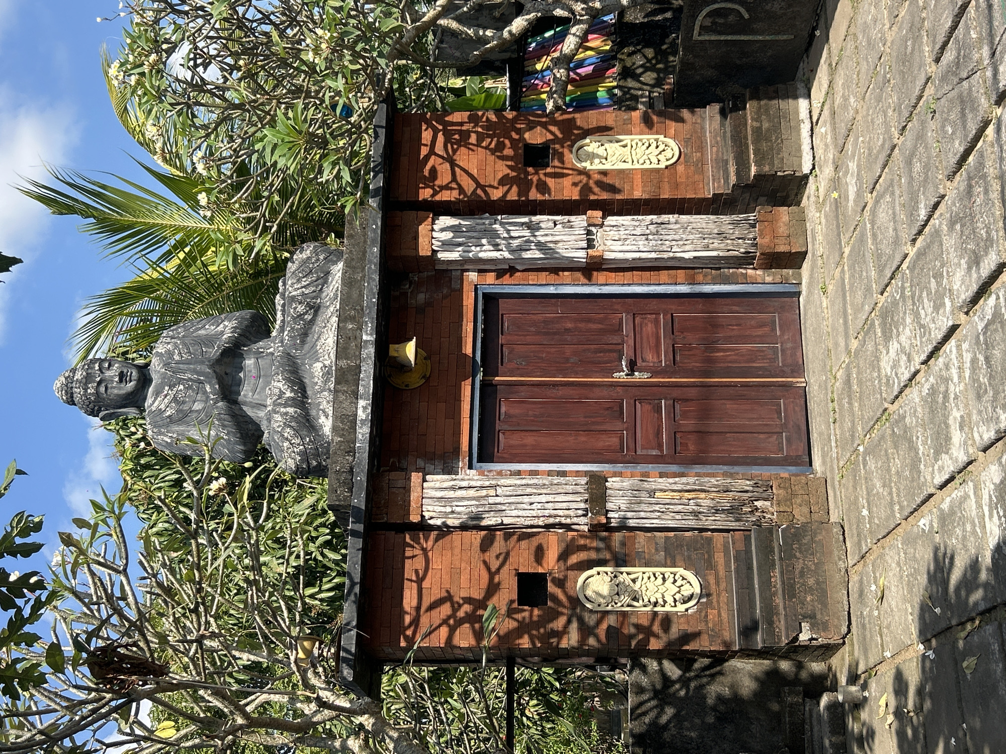 A stone statue of the Buddha is perched atop a brick structure with a double wooden door in Mengwi, Indonesia. The statue is in a meditative pose. The brick structure is adorned with two carved decorative panels on either side of the door. Several trees with lush green foliage surround the structure, and palm fronds are visible in the background. The ground in front of the structure is paved with stone slabs, and fallen leaves and small white flowers are scattered on the paving. The sky above is blue with a few scattered clouds. The overall scene suggests a serene and spiritual atmosphere.