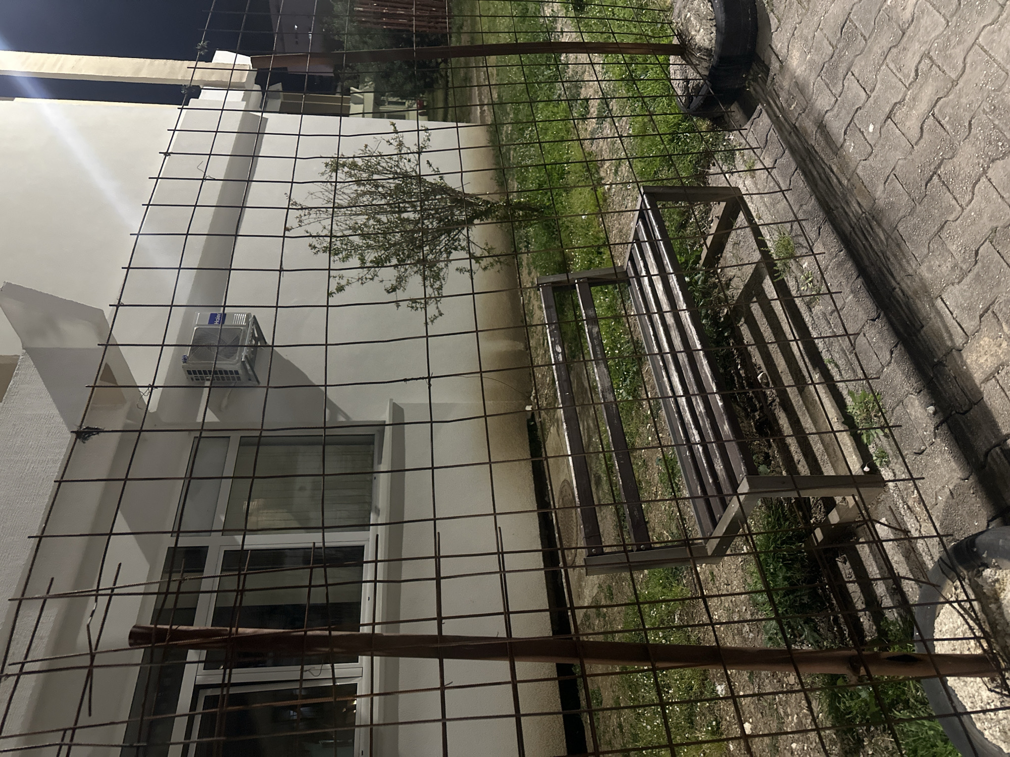 The image shows an outdoor scene with a metal wire fence in the foreground. Behind the fence is a modern, multi-story building with white walls. A window and an air conditioning unit are visible on the building's facade. A small tree is located near the building. A metal bench sits on a patch of grass between the fence and the building. To the right, paving stones are visible on the ground. The scene appears to be in the city of Budva, Montenegro, due to the style of architecture.