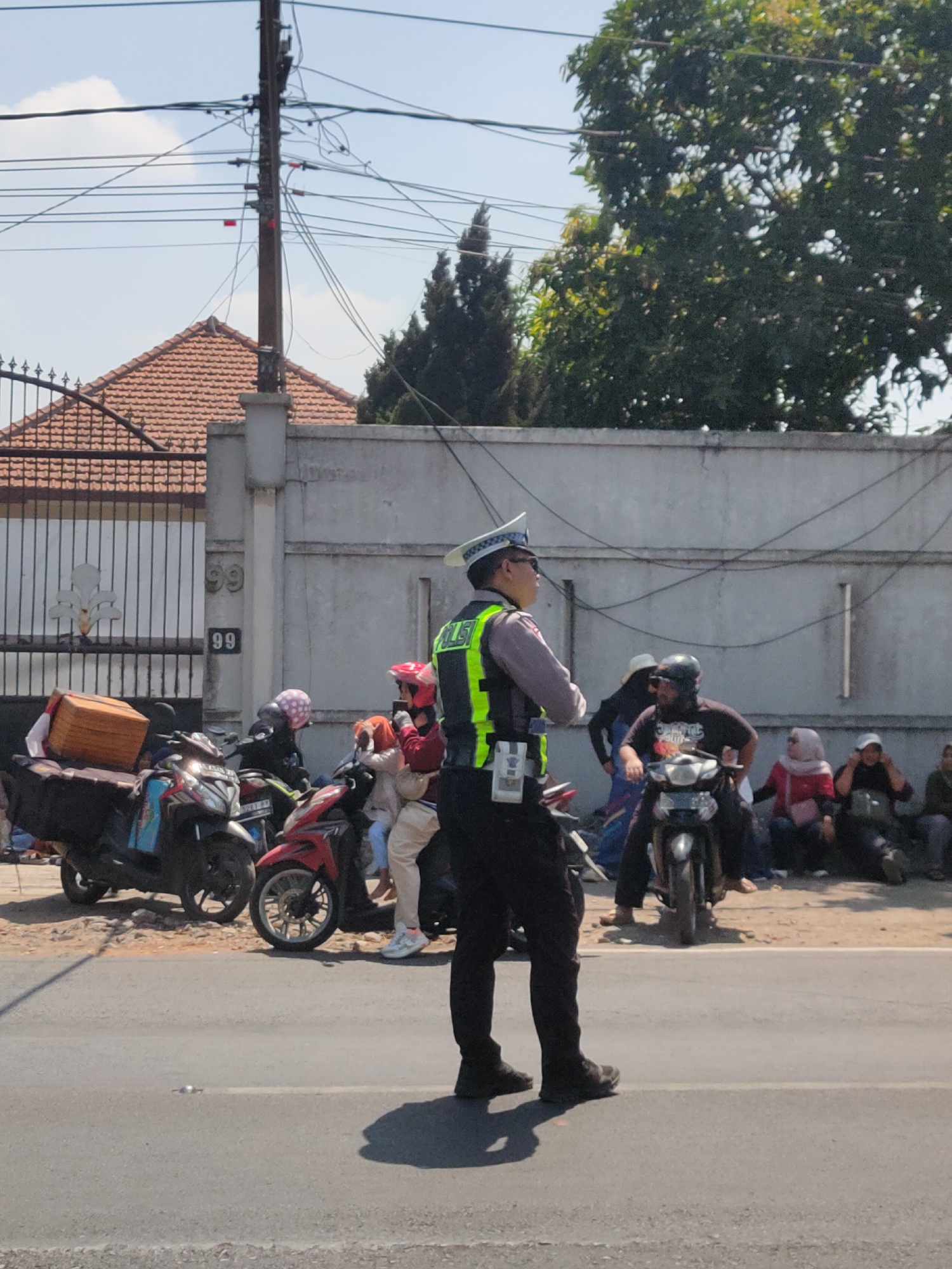 In Ngasem, Indonesia, a traffic police officer stands by the roadside, directing traffic. He wears a reflective vest over a gray uniform and a white and blue cap. Several motorcycles are parked or moving on the asphalt road. A few people are seated on the side of the road, some with motorcycles. Behind them, a gray concrete wall runs along the street, with a metal gate and a tiled roof house in the background. Trees and power lines are visible against a clear blue sky.