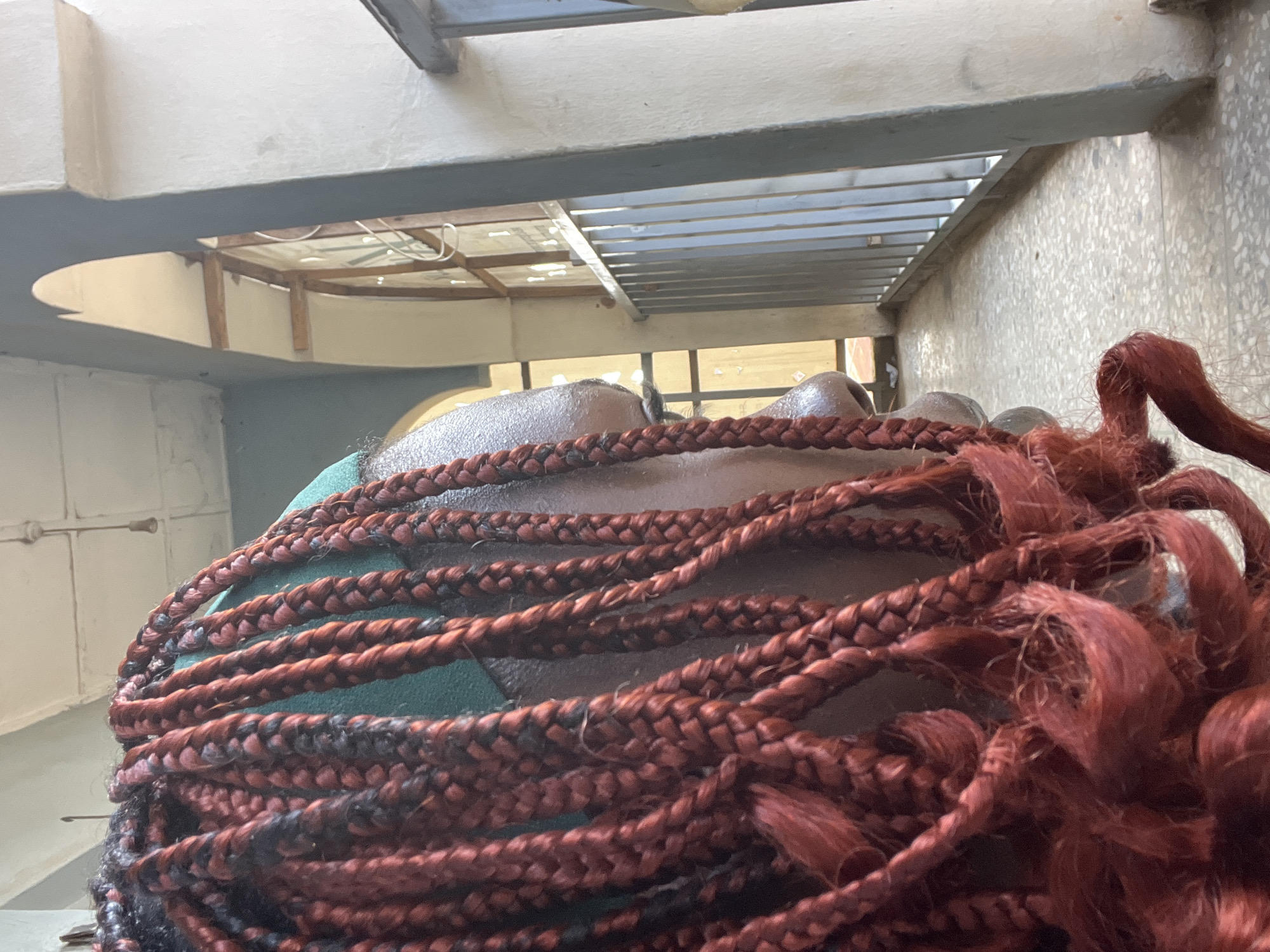 This image depicts a close-up view of a person's head and braids, with their face turned to the right. The person is wearing a green head covering. The braids are a mix of red and black. In the background, there is a balcony or walkway with a metal railing and an arched opening leading to what appears to be a building. The flooring is tiled. The scene is outdoors, with sunlight casting shadows. The overall environment suggests an urban setting, possibly in the city of Jos, Nigeria, given the architectural style and natural lighting.