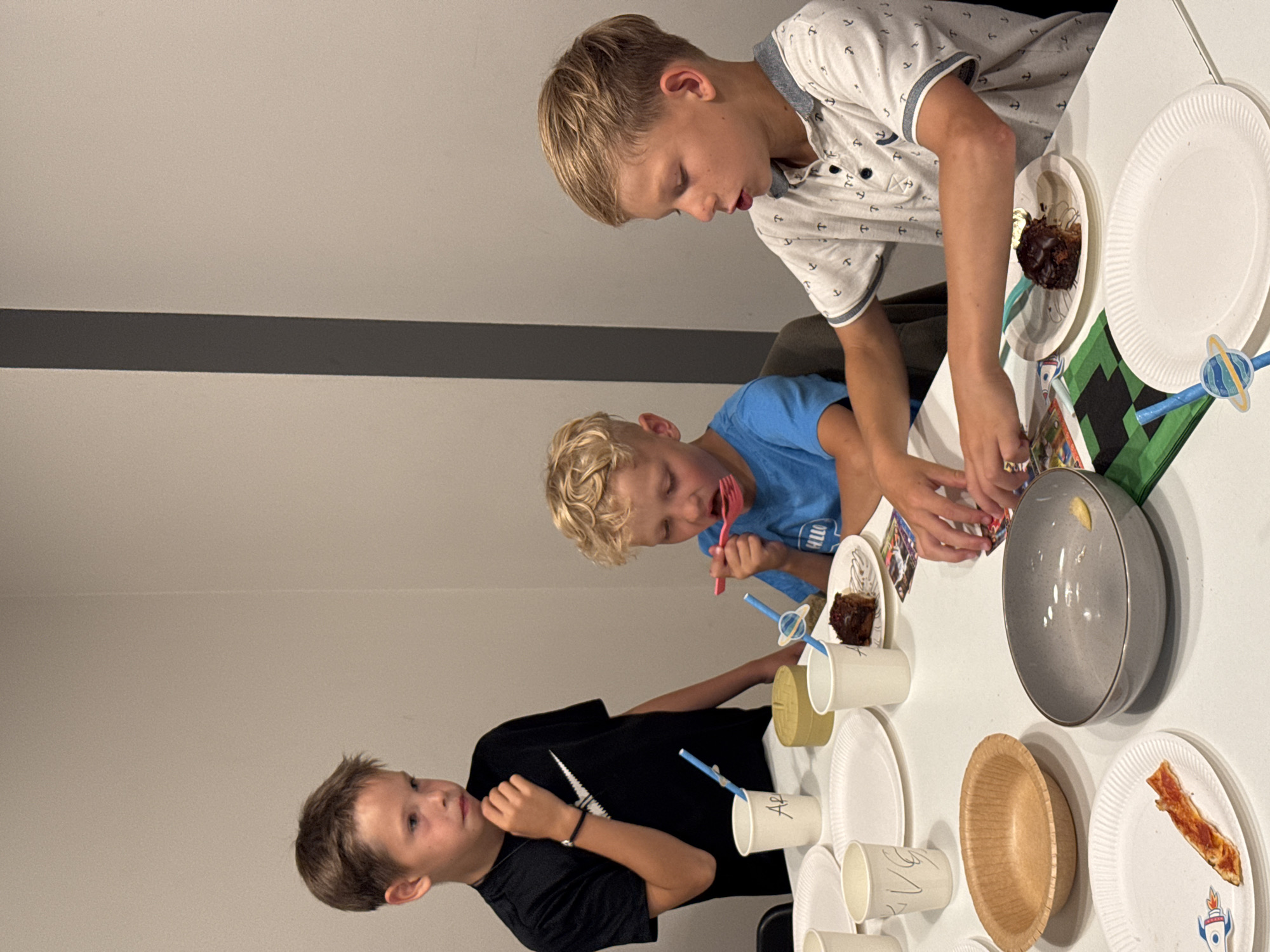 The image depicts three young boys at a table, engaged in what appears to be a birthday party or a similar celebration. The setting is indoors, with a plain, light-colored wall in the background featuring a dark horizontal stripe. The table is covered with white paper plates, cups, and some food items, including what looks like a piece of cake and a slice of pizza. One boy, wearing a white polo shirt with anchor patterns, leans over the table, looking intently at something. Another boy, in a blue t-shirt, is actively eating with a pink fork. The third boy, in a black t-shirt, is also seated at the table, looking away from the camera. Some of the paper cups have handwritten names on them, one clearly legible as "Apu". There are also decorative elements on the table, including a green and black checkered item and a straw with a planet-shaped decoration. The overall mood seems casual and focused on the activity at hand, suggesting a child's gathering. The lighting suggests it could be daytime, possibly indoors with artificial lighting supplementing natural light. The location is indicated as Riga, Latvia.