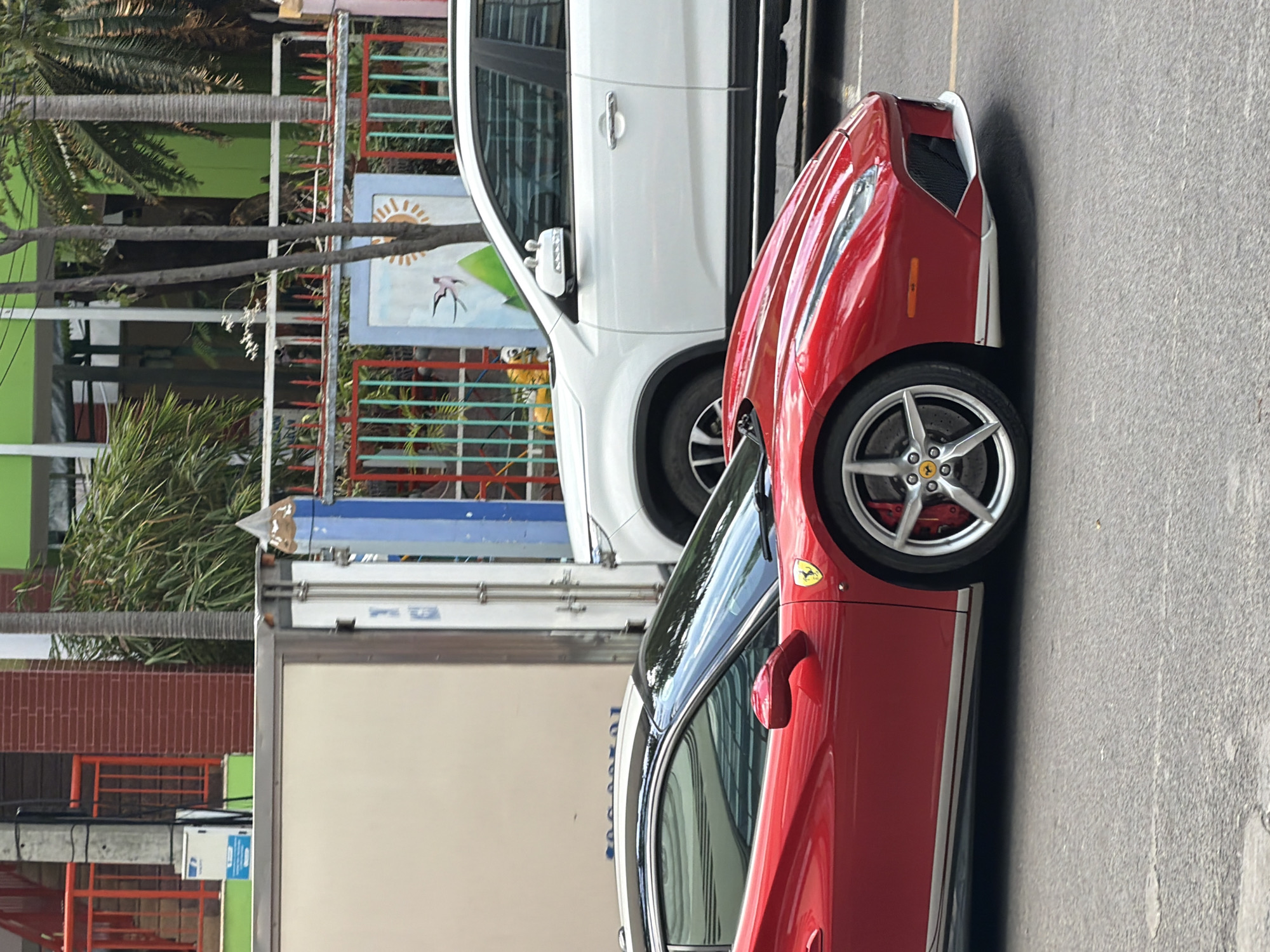 The image shows a red Ferrari sports car and a white SUV parked on a city street. The Ferrari is in the foreground, with its side profile clearly visible. The white SUV is behind it. In the background, there is a building with a green facade, a fence with colorful metal bars, and some trees. The scene appears to be in an urban environment in Nha Trang, Vietnam, as indicated by the presence of typical street infrastructure and vegetation. There are no people visible in the image. The weather seems to be overcast, as there are no harsh shadows.
