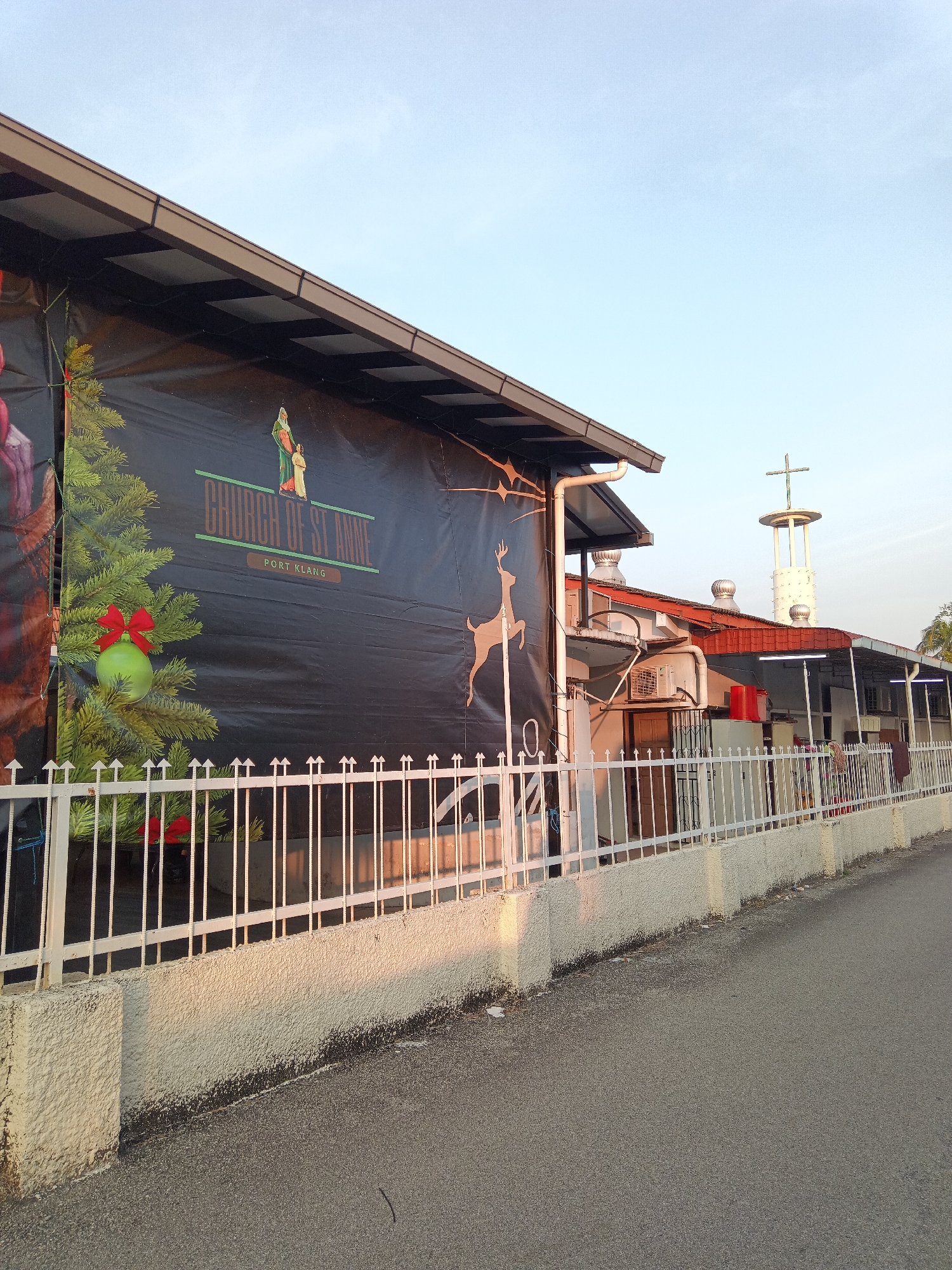 A long shot of the Church of St Anne, Port Klang, Malaysia is captured during daylight hours. The church building is a modest structure with a dark-colored banner adorning its facade. The banner features festive Christmas imagery, including a decorated Christmas tree with ornaments and a depiction of the Holy Family. The words "CHURCH OF ST ANNE" are prominently displayed on the banner in a green font, with "PORT KLANG" in a smaller font below. A white metal fence separates the church property from the paved road in the foreground. The sky is clear and blue, indicating fair weather. In the background, a bell tower with a cross at its apex is visible, a characteristic feature of many churches. The overall scene suggests a peaceful and serene atmosphere, likely capturing a moment of community gathering or religious observance.