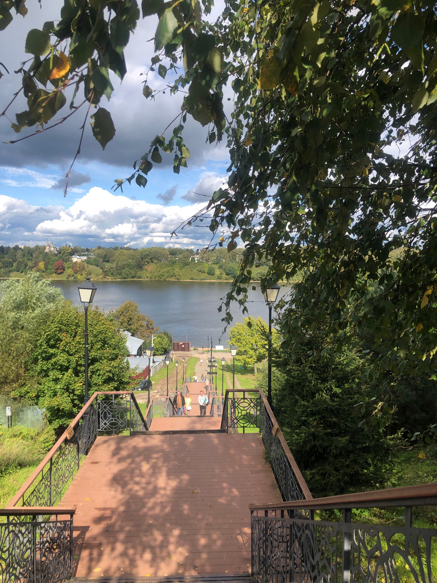 The image captures a view of a wide wooden staircase with ornate metal railings leading down towards a body of water. The staircase is lined with lampposts. Several people are seen walking down the stairs, some in groups. Beyond the staircase, a river stretches across the landscape, with lush green trees and buildings dotting the opposite bank. A church with a dome is visible in the distance on the far bank. The sky is partly cloudy, with patches of blue. The scene appears to be set in Tutaev, Russia, a city known for its riverside scenery.
