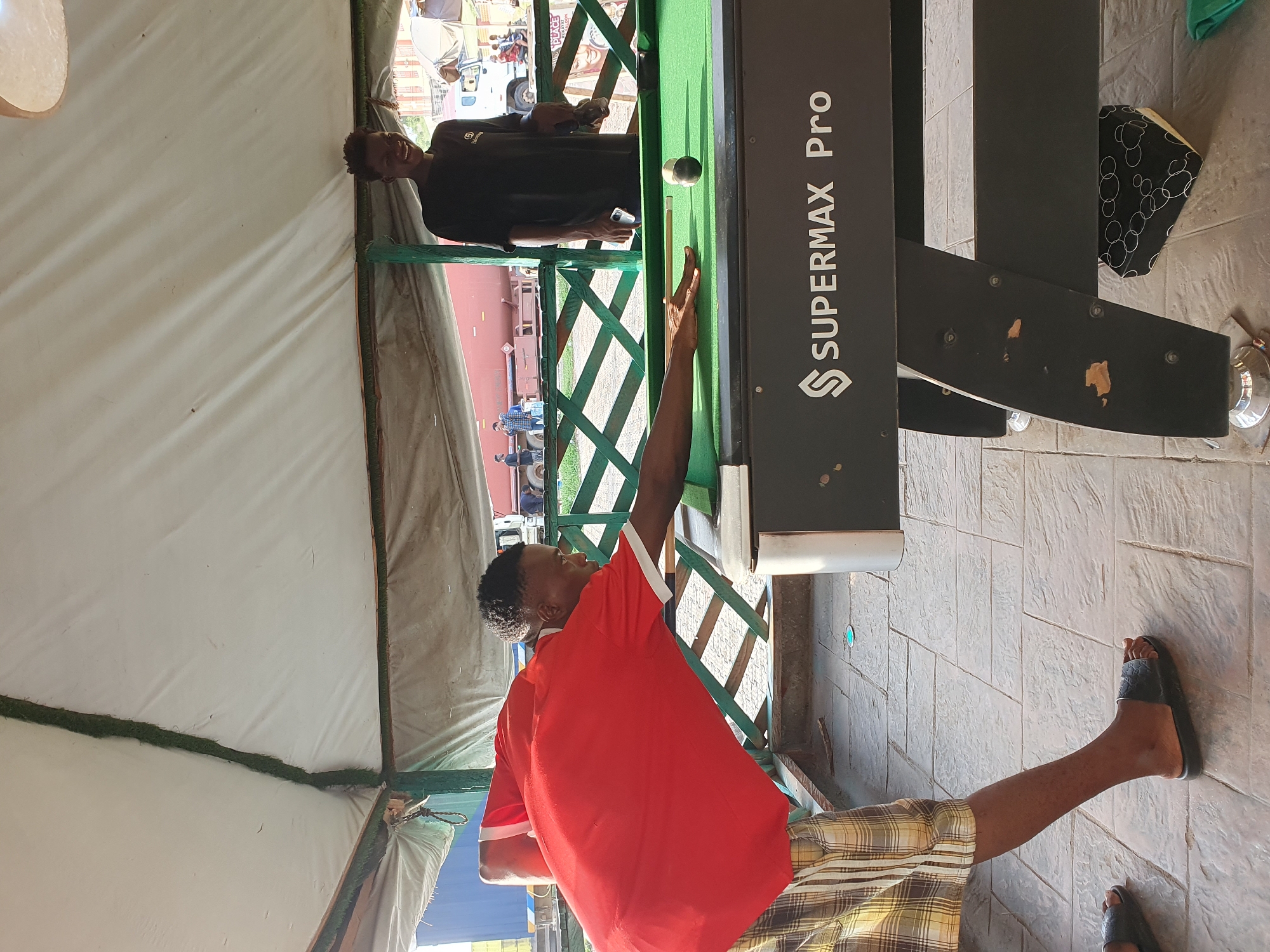 Two young Black men are playing billiards under a white tarp canopy in Lokoja, Nigeria. The man in the foreground, wearing a red t-shirt and plaid shorts, is bent over the green billiard table, aiming his cue stick at a white ball. His left hand is on the table for stability. The other man, dressed in a black t-shirt, stands behind him, smiling and holding a smartphone. The billiard table has the "SUPERMAX Pro" logo on its side. The background shows a street scene with vehicles and buildings, suggesting an urban outdoor setting. The ground is paved with interlocking stones.