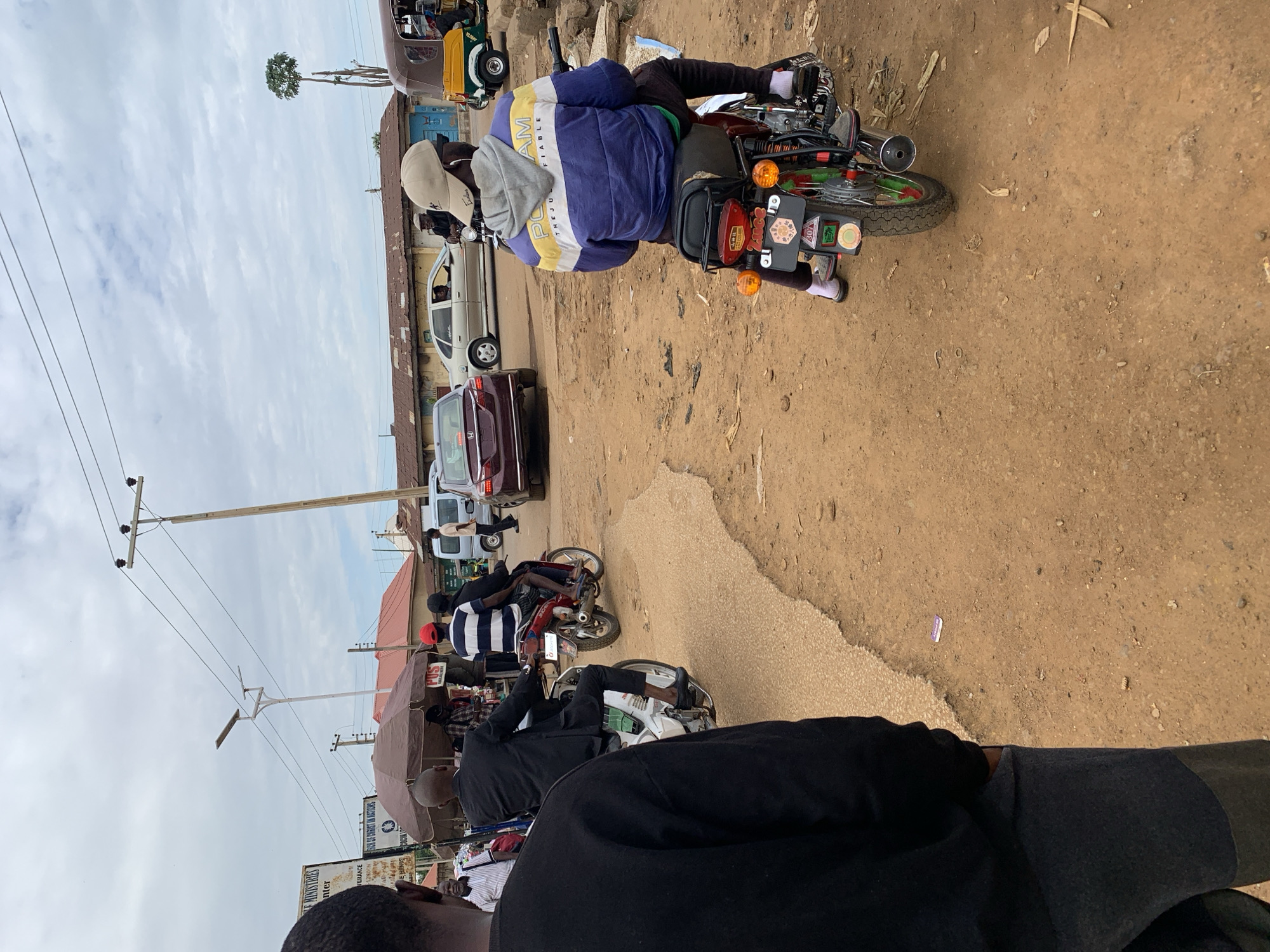 This image depicts a bustling street scene in Rafin Par, Nigeria, characterized by both paved and unpaved sections, vehicular traffic, and commercial activity under an overcast sky.
In the foreground, the back of a person wearing a dark long-sleeved shirt and dark grey sweatpants is visible on the left, standing on a reddish-brown dirt shoulder. A small purple wrapper lies on the dirt ground to their right.
Moving towards the mid-ground, a slightly elevated, darker asphalt road snakes through the dirt. Several individuals are operating or riding on motorcycles. On the left, a man in a black suit with his lower legs bare, wearing black dress shoes, is astride a white motorcycle (possibly a Honda Cub clone) and moving away from the viewer. Behind him and slightly to the right, two people are on a red motorcycle; the passenger, wearing a red cap and a red and white striped shirt, is facing away.
On the right side of the paved section, another man, wearing a blue, yellow, and white hooded jacket, a light beige cap, dark pants, and white socks with open-toed sandals, is riding a red motorcycle. The motorcycle's rear is prominently visible, displaying a license plate with "TARABA" and "307Y" along with other stickers.
Vehicular traffic includes a maroon sedan (likely a Honda) on the left lane, and a silver sedan (possibly a Volkswagen) further back. On the far right, a brown and yellow motorized tricycle taxi, commonly known as a Keke Napep, is parked or in motion, with a white van or mini-bus behind it.
The background features several single-story buildings with corrugated metal roofs, some painted reddish-brown and others silver. A prominent brown market umbrella stands on the left, sheltering a stall where several people are gathered, and a "POS" sign is visible. Another sign further left partially reads "CHURCH OF CHRIST IN NATIONS" and "MINISTRIES." Overhead, multiple wooden utility poles support power lines that crisscross the sky. A tall white pole with a solar street light fixture extends upwards from the left side of the street. On the far right, a very tall, slender tree with sparse foliage stands against the cloudy sky. The ground throughout the unpaved areas is a reddish-brown dirt, with minor debris and scattered plant material.