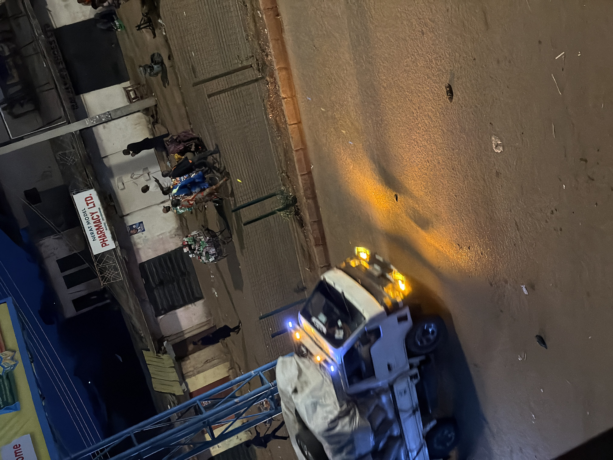 In Bukuru, Nigeria, at night, a flatbed truck with its headlights illuminated is parked on a street. The truck is covered with a large, light-colored tarp. Several people are visible on the sidewalk in the background, some standing and others appearing to be walking or pushing a cart. The buildings in the background appear to be shops or residences, with some signs visible, including one that reads "NERAT MODEL PHARMACY LTD." The street is paved and appears somewhat worn. The lighting suggests an urban environment at night, with artificial lights from the truck and buildings illuminating the scene.