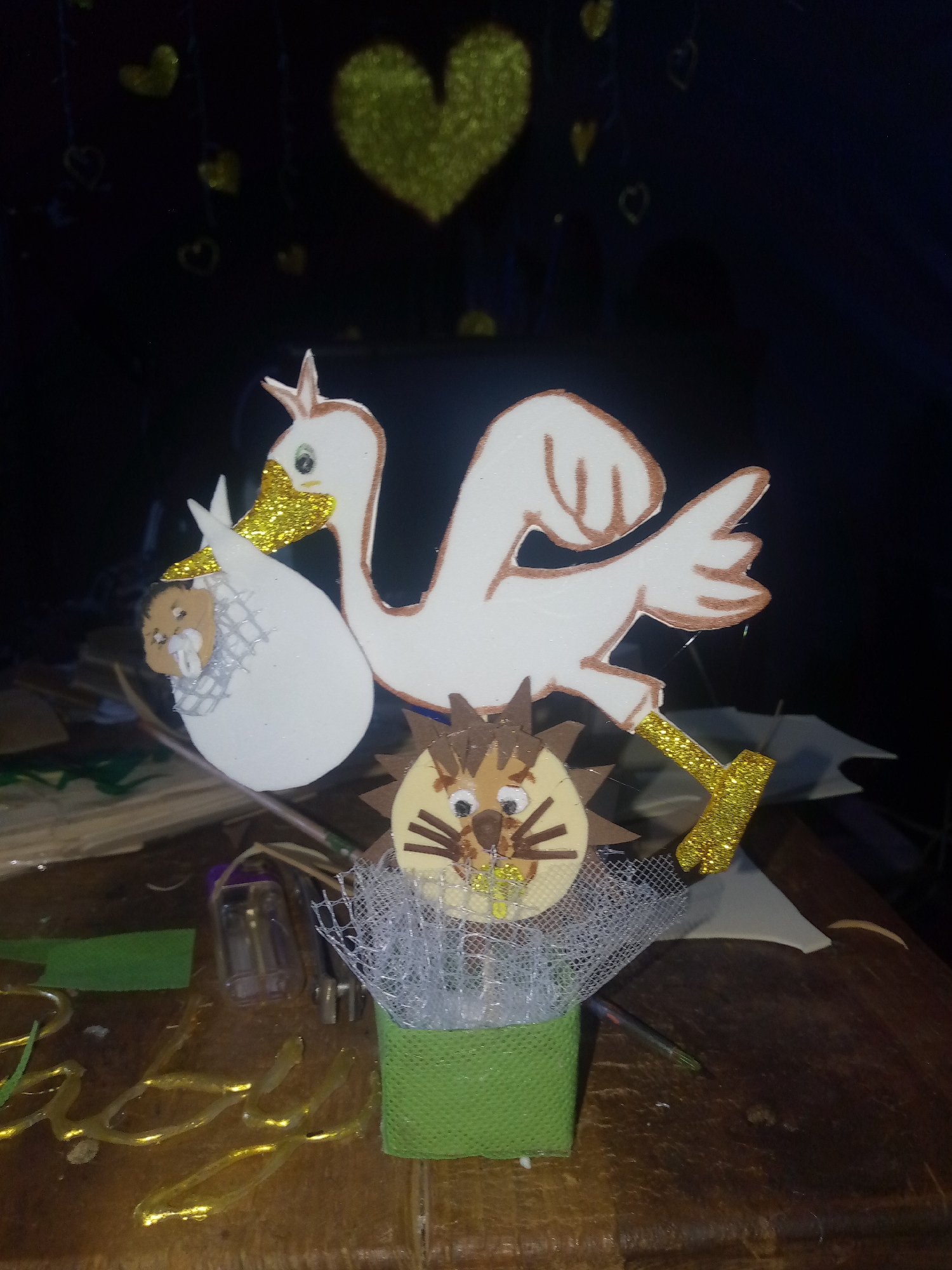 The image depicts a handcrafted decoration featuring a stork carrying a baby in a sling and a lion's head emerging from a green square base covered in white netting. The stork and lion figures are made of painted craft materials with glitter accents. In the background, out-of-focus gold heart-shaped decorations hang against a dark backdrop. The decoration is situated on a wooden surface among various craft supplies. There is no visible information to ascertain that the location is in Juan Griego, Venezuela.