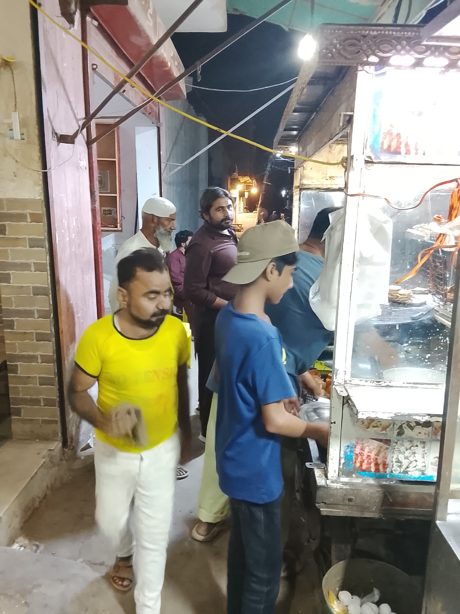The image captures a bustling nighttime street scene, likely centered around a food stall. Multiple men and a boy are present, suggesting a common gathering spot for food.
In the foreground, a man in a vibrant yellow t-shirt, white pants, and sandals, with a dark beard and mustache, walks towards the left, appearing slightly blurred due to motion. His expression is neutral. To his right, a young boy wearing a blue t-shirt and a tan baseball cap, which has "Columbia" visibly printed on it, stands facing a food stall, his head slightly tilted and a faint smile on his face. He appears to be holding a metallic bowl or utensil.
Behind them and to the right is a food stall with a glass display cabinet. Inside the cabinet, various packaged food items or ingredients are visible, including what looks like small, flat, round items (possibly crackers or bread) and colorful snack bags. At the bottom right of the stall, a bucket filled with white eggs is placed on the ground. A man in a dark blue shirt, seen from the back, is actively working at the stall, leaning over to prepare food. Another white bag hangs near him.
Further in the background, several other men are visible. A man wearing a white skullcap (kufi) and a white shirt with a long white beard looks towards the left. Beside him, a man with a beard and mustache, dressed in a dark reddish-brown shirt, looks to the right. A younger man in a maroon shirt is also visible behind them.
The setting appears to be an urban or semi-urban area at night, illuminated by a bare light bulb hanging above the food stall and other distant lights in the dark background. Yellow and black electrical wires crisscross the upper part of the frame. On the left, a brick wall and a reddish-pink door or shutter of a building are visible, adding to the streetscape ambiance. The ground is uneven, suggesting an unpaved or worn path.