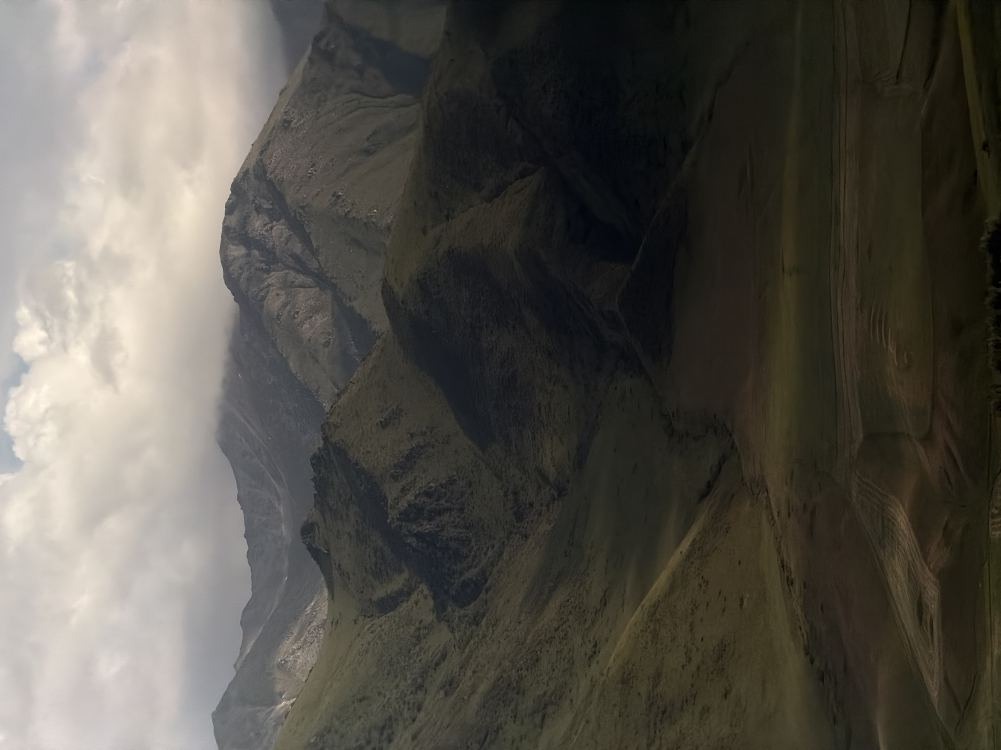 This is a still, vertically oriented, wide-angle photograph of a mountainous landscape. The scene is dominated by steep, green slopes of grass and rock, with distinct ridges and valleys carved into them. The lighting suggests it is daytime, with clouds obscuring parts of the sky and casting shadows on the mountainside. The overall mood is one of natural grandeur and ruggedness. The image is a landscape scene, with no people or discernible objects other than the natural topography. The location is Karagai Bulak, Kyrgyzstan, a region known for its dramatic mountain terrain. The weather appears to be mixed, with sunshine breaking through cloudy skies. There is no visible text in the image.