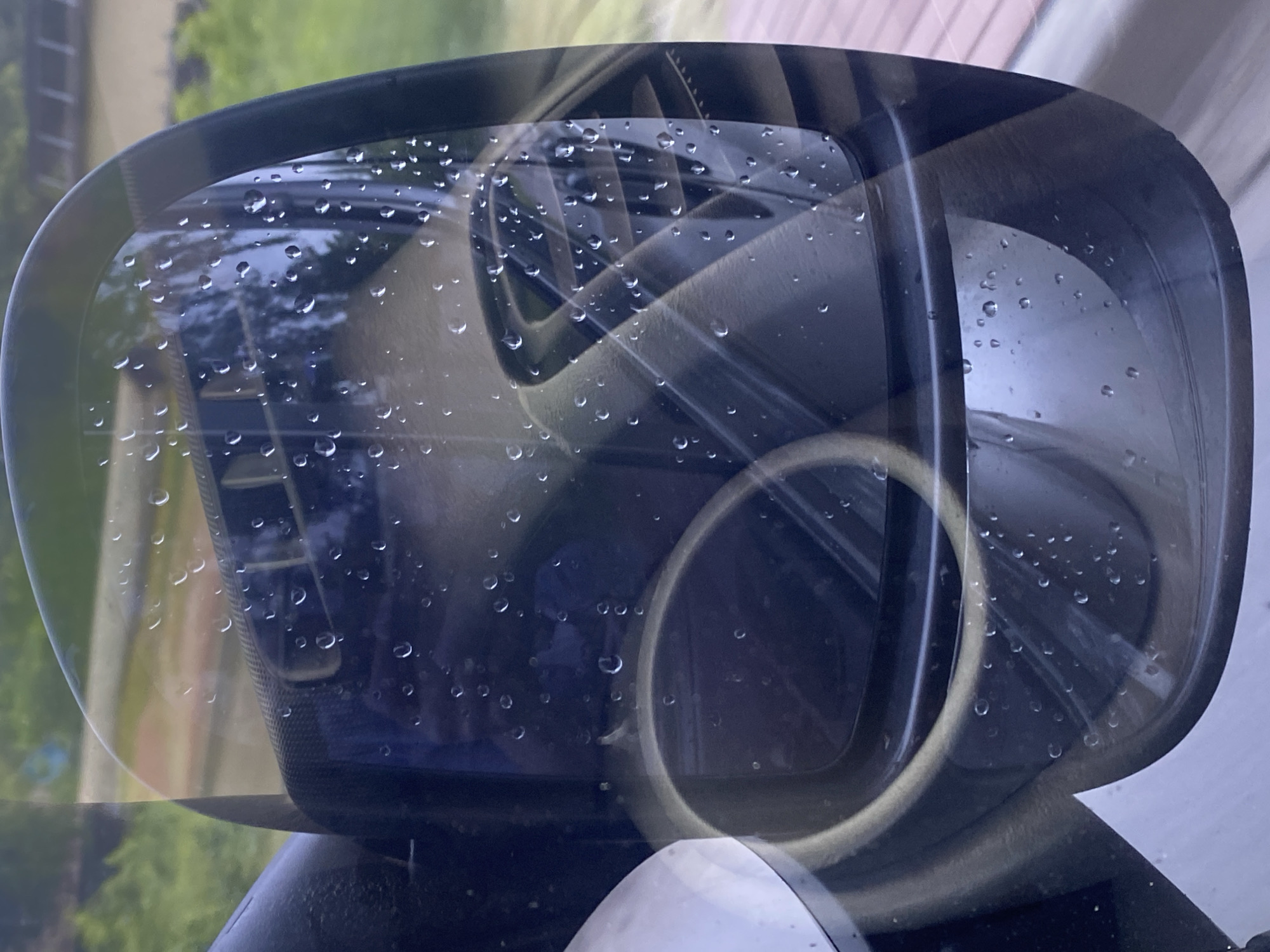 The image is a close-up, angled shot of a car's side mirror, reflecting the interior dashboard. The mirror's surface is covered with numerous water droplets, suggesting it is raining or has recently rained. The reflection shows part of the car's dashboard, including air vents and a circular vent or dial. The exterior environment reflected in the upper portion of the mirror appears to be a green, leafy area with some structures in the background, possibly a road or walkway. The overall impression is that of a vehicle parked outdoors in Pukhivka, Ukraine, during inclement weather.