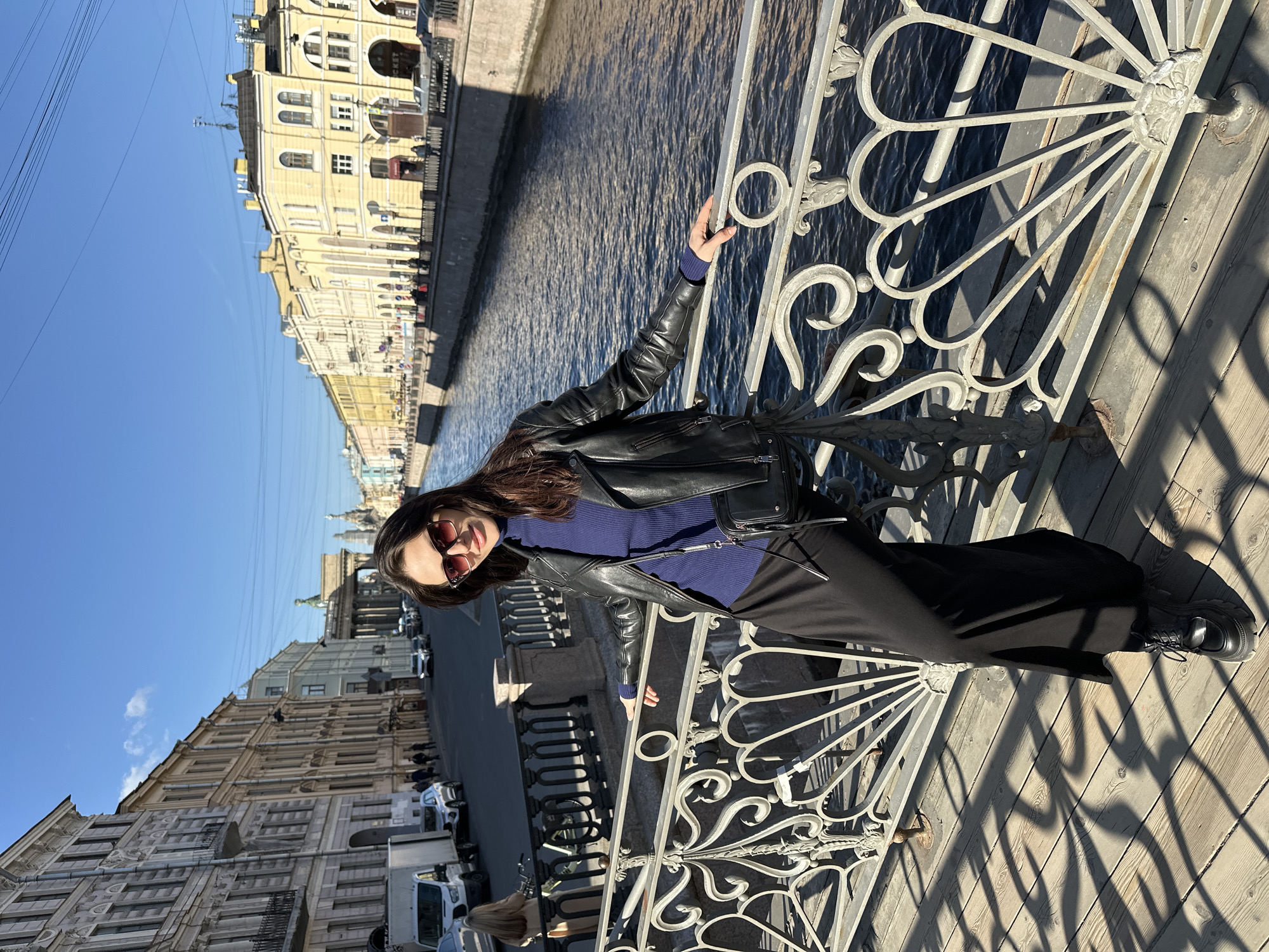 A woman poses for a photograph on a sunny day in Saint Petersburg, Russia. The image, rotated 90 degrees clockwise, depicts her standing on a wooden bridge, leaning against an ornate, light-colored metal railing that features intricate scrollwork and fan-like patterns. She has long, dark hair and wears dark sunglasses, a dark blue turtleneck or sweater, a black leather jacket, black wide-leg pants or a skirt, and black chunky boots. Her left hand rests casually on the bridge railing, and she looks towards the camera with a slight smile.
To her right, the deep blue water of a canal flows, reflecting the bright sky. Along the canal banks, rows of classical-style buildings with intricate facades, arched windows, and decorative elements stretch into the distance. Most buildings are painted in shades of light yellow, beige, or grey. Cars are parked along the streets flanking the canal. Overhead, against a clear, bright blue sky with a few wisps of clouds, utility lines are visible. The sun casts long, distinct shadows from the bridge railing onto the wooden planks, suggesting mid-morning or afternoon. The architecture strongly indicates the setting is Saint Petersburg. No discernible text is visible on buildings or signs.