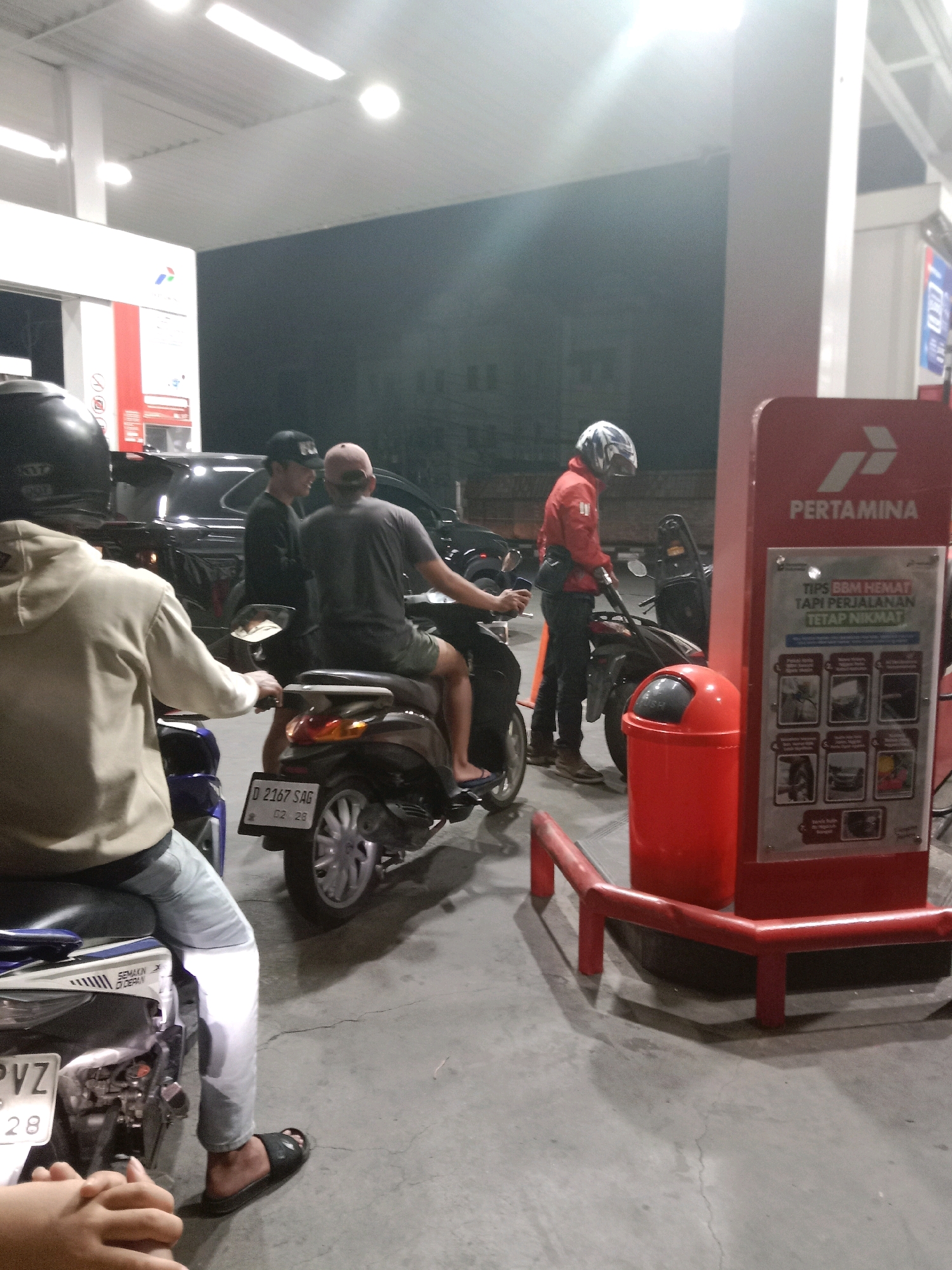 The image captures a scene at a Pertamina gas station in South Jakarta, Indonesia, likely at night. In the foreground, a person wearing a light-colored hoodie and jeans is seated on a motorcycle, facing away from the camera. Their hands are visible in the lower left corner, resting on the motorcycle's seat. To the right, another individual in a dark t-shirt and shorts is also seated on a motorcycle, with a license plate reading "D 2167 SAG".
Further back, a person in a red jacket and helmet is operating a fuel pump, with the nozzle extended towards a motorcycle. Another individual wearing a black baseball cap and dark clothing stands near the fuel pump. A black SUV is parked behind the people on the motorcycles, partially obscured. The gas station features typical amenities like a large red fuel dispenser with Pertamina branding and informational posters. A red trash can is positioned near the dispenser. The environment appears to be an outdoor, paved area, illuminated by overhead lighting, creating shadows on the ground.