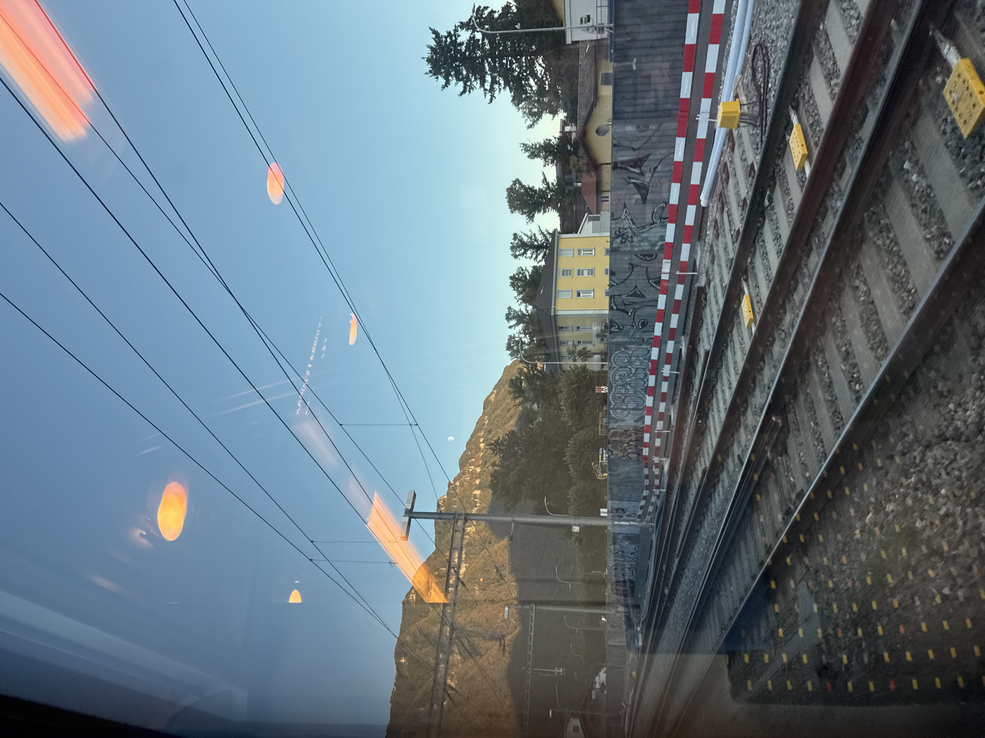 This is a photograph taken from a train window, looking out at a railway track and the surrounding environment in the city of Val Mara, Switzerland. The time of day appears to be late afternoon or early evening, with the sun casting long shadows.
The foreground is dominated by multiple sets of parallel railway tracks. The tracks are laid on a bed of gravel, and some have yellow markers along the sides. A reflective surface, likely the train window, distorts the view, creating reflections of orange lights and overhead power lines.
Beyond the tracks, a concrete wall covered in graffiti runs horizontally across the image. This wall is topped with a red and white striped barrier. In the background, there are several buildings, including a prominent yellow building with multiple windows. Trees are scattered throughout the scene, and in the distance, a large mountain range is visible. A sliver of the moon can be seen in the clear blue sky.
There are no people visible in the image. The focus is on the infrastructure of the railway and the landscape. The overall impression is one of movement and observation from a moving vehicle, with the natural environment of the mountains contrasting with the man-made elements of the railway and buildings.