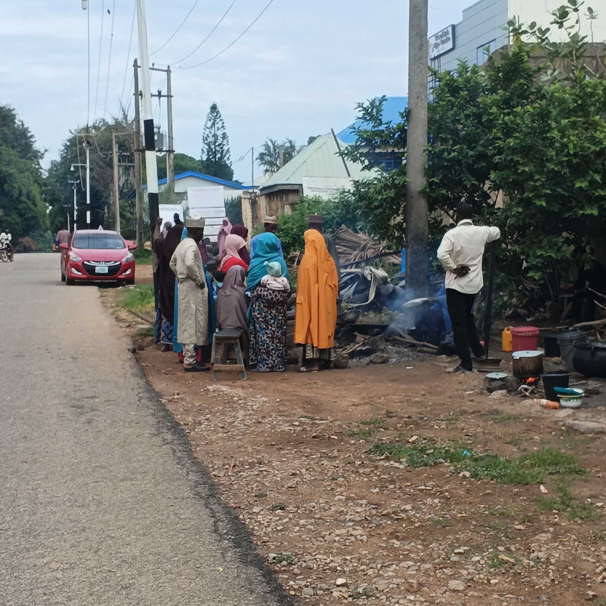 In Unguwar Rimi, Nigeria, a street scene unfolds with a red sedan parked on the side of an asphalt road. A group of people, predominantly women and children dressed in traditional attire, are gathered on the dirt shoulder of the road. Some are standing, while a child sits on a wooden stool. A man in a white shirt and black pants stands near a cooking fire with several pots. The environment includes utility poles, overhead wires, lush green trees, and buildings in the background, one with a blue roof. The ground is a mix of dirt, gravel, and sparse green vegetation.