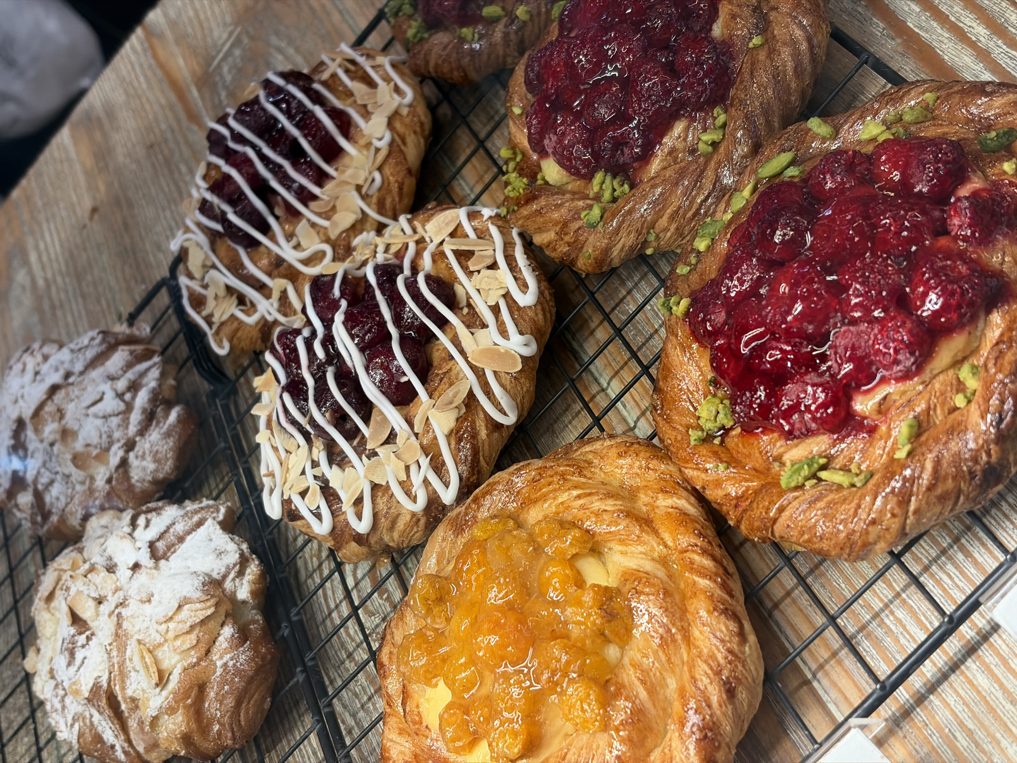 A high-angle, close-up shot displays a variety of fresh pastries arranged on a black wire cooling rack, set against a light-colored wooden surface. The scene captures a selection of sweet baked goods, likely danishes and almond croissants, highlighting their textures and toppings.
In the upper right and middle right, there are two distinct raspberry danishes. These feature a braided or twisted golden-brown pastry base, generously topped with glossy, bright red raspberries and a scattering of vibrant green pistachio pieces. Another similar raspberry danish is partially visible in the top left corner, out of focus.
Towards the center and upper left, two more danishes are visible, distinguished by a dark red, berry-like filling (possibly cherry or mixed berry) and decorated with intricate white icing drizzles and delicate slivered almonds. These pastries appear to have a more rectangular or square shape.
In the bottom center, a round danish stands out with a bright yellow-orange fruit filling, possibly apricot or peach preserves, spread over a creamy base. Its pastry is also a rich golden-brown.
On the left side of the cooling rack, two almond croissants or similar pastries are heavily dusted with white powdered sugar and adorned with slivered almonds. The one in the foreground is fully visible, while the one behind it is partially obscured.
The wooden surface beneath the cooling rack shows distinct grain patterns, suggesting a natural wood table or counter. The overall lighting is bright and warm, emphasizing the appealing golden-brown crusts and vibrant fillings of the pastries. There is no visible text in the image. The setting suggests a bakery or cafe, in Saint Petersburg, Russia.