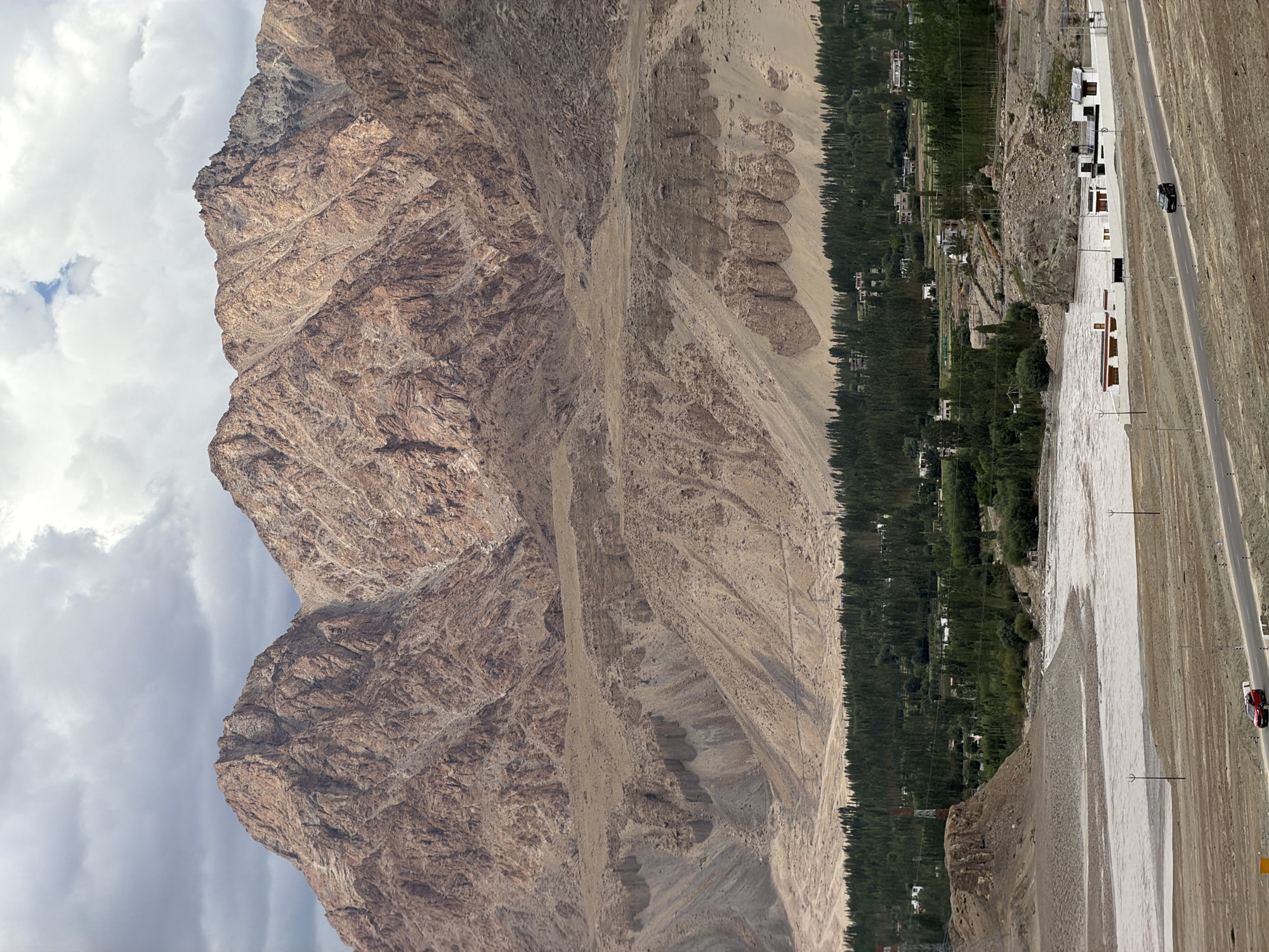 The image shows a vast, arid mountain landscape with a settlement nestled in a valley. The mountains, characterized by their rugged, eroded surfaces and shades of brown and tan, dominate the upper portion of the frame. Below the mountains, a barren, sandy slope transitions into a verdant valley. This valley is densely populated with tall, dark green trees, interspersed with small, white buildings that form a settlement. A wide, shallow river, appearing milky or sandy, flows through the foreground, separating the settlement from a road. The road, a dark asphalt strip, runs diagonally across the lower right quadrant of the image. Two vehicles are visible on the road: a red car is parked on the shoulder, and a black car is in motion. The sky above is overcast with dramatic, dark gray clouds, hinting at a possibly unsettled weather condition. The scene conveys a sense of isolation and the stark beauty of a high-altitude desert environment.