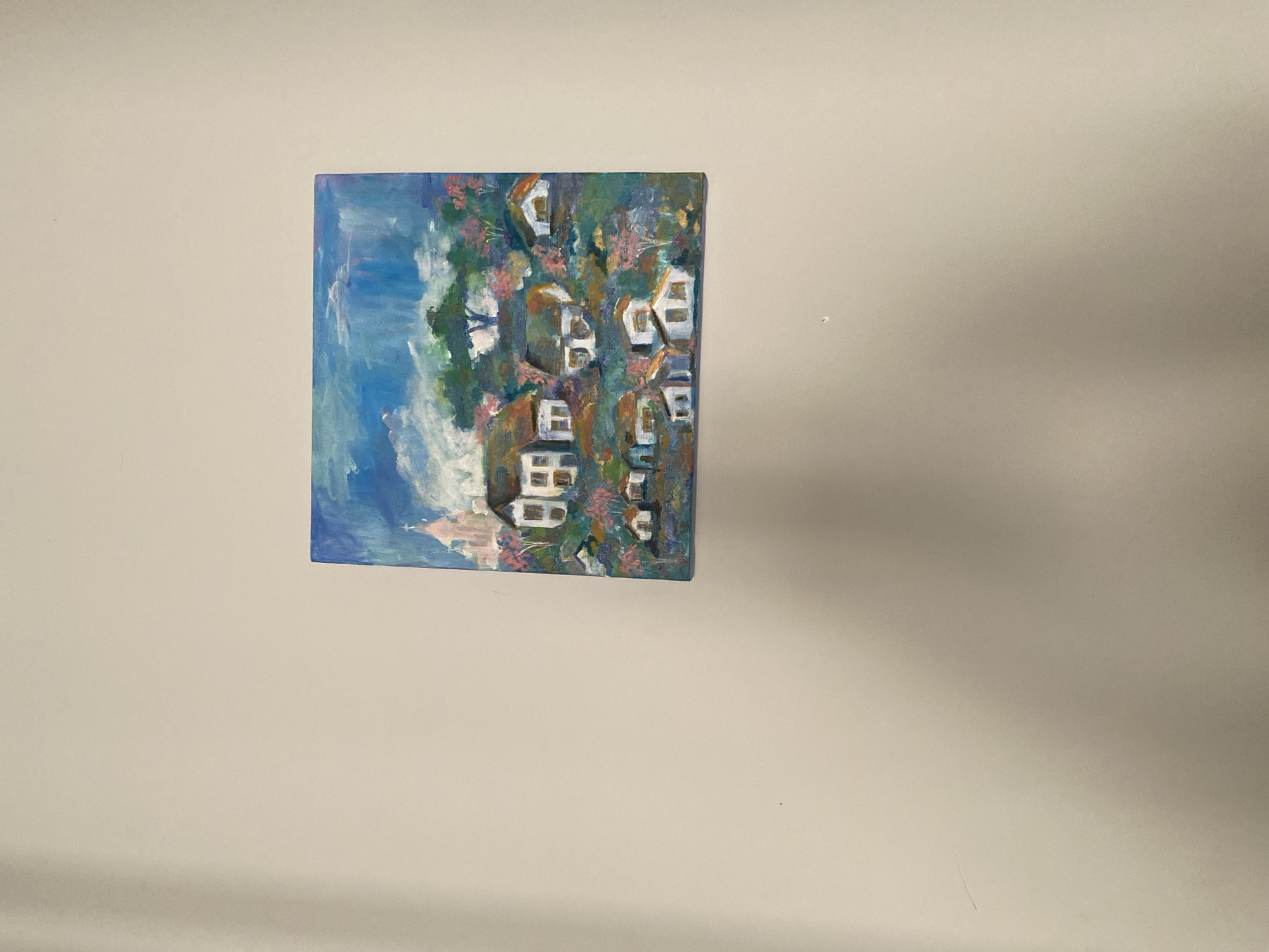 An oil painting hangs on a neutral-colored wall. The painting depicts a cluster of houses against a blue sky and green foliage, evoking a scene similar to the architecture found in hillside towns. The houses are rendered with white facades and simple square or rectangular windows. The brushstrokes are visible, giving the artwork a textured and somewhat impressionistic feel. The environment consists of blue sky with splashes of white, and patches of pink that imply foliage. The scene does not provide enough information to pinpoint a precise location in Lâm Viên - Đà Lạt, Vietnam.