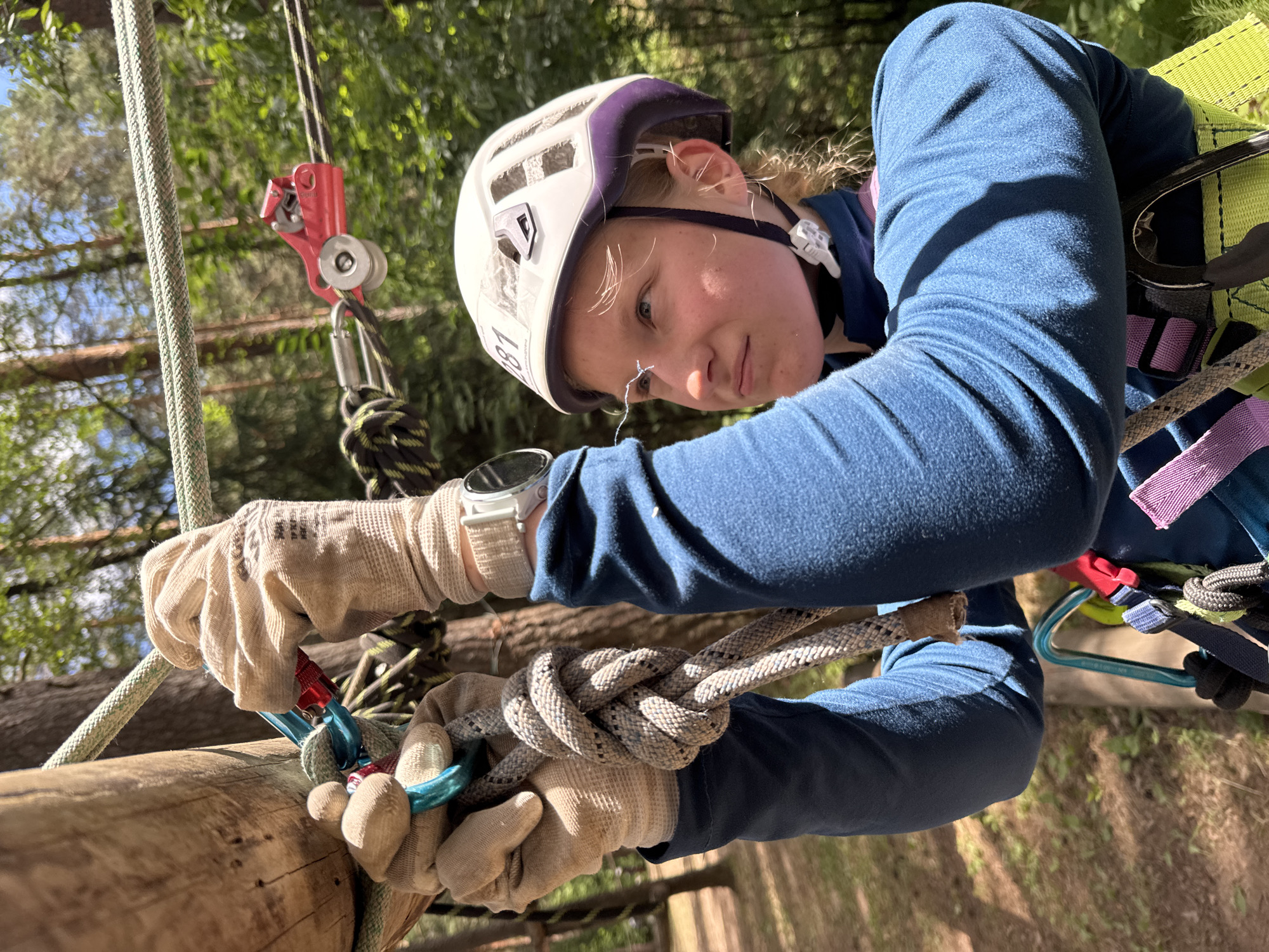A young woman, wearing a white and purple helmet, a blue long-sleeved shirt, and a green and yellow harness, is seen from the chest up while participating in an outdoor ropes course. She is wearing beige gloves and appears to be securing a thick, grey rope with a knot. Her left hand is positioned above her right. A portion of a thick wooden pole is visible on the left side of the frame. Behind her, a red pulley system is attached to a horizontal rope. The background consists of lush green trees and a bright blue sky, indicating an outdoor environment, likely a park or adventure attraction. The image is taken in Chaikovsky, Russia.