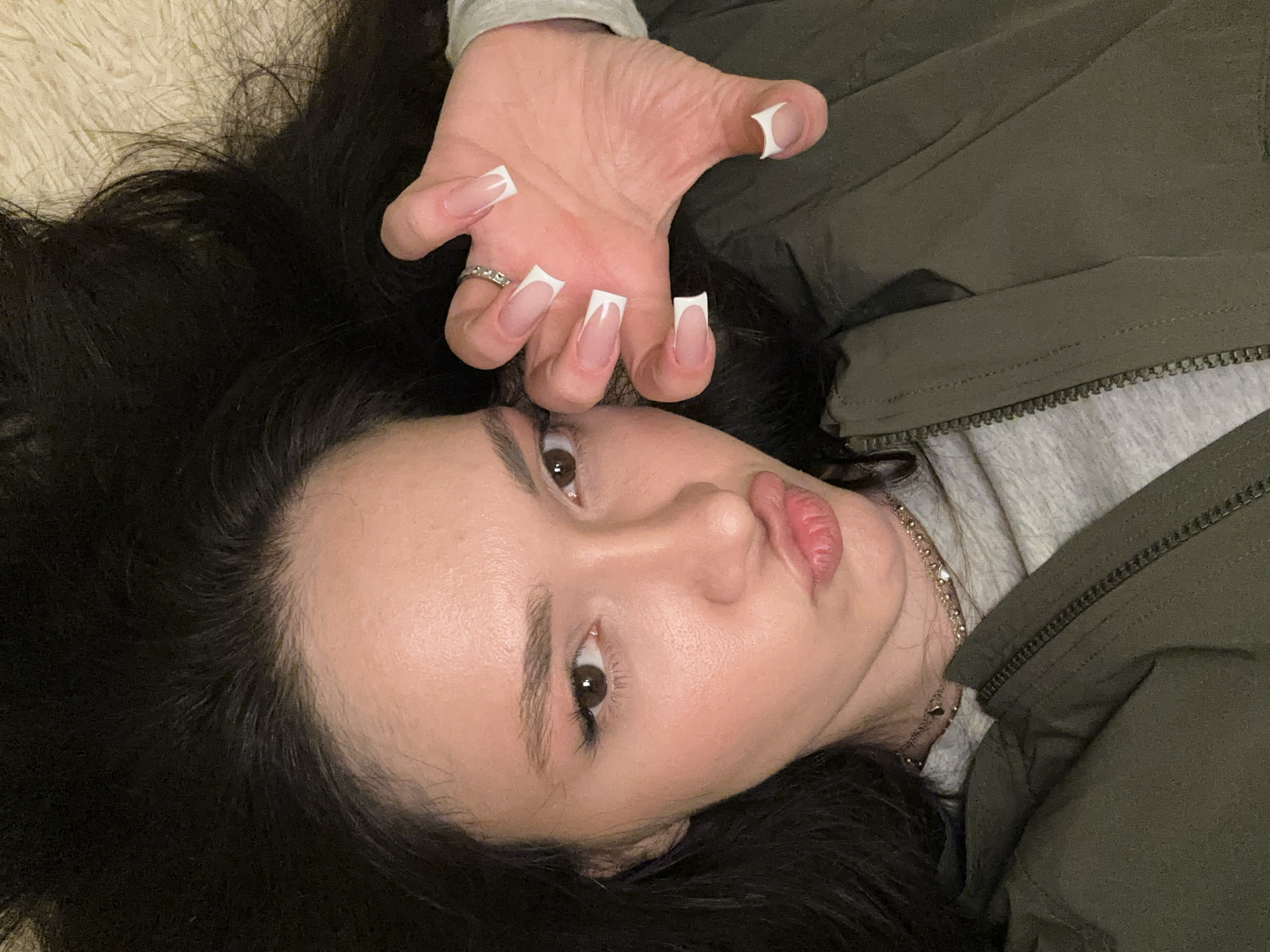 A close-up image depicts a young adult female, positioned horizontally, possibly lying on a light-colored, fuzzy or textured surface. Her dark hair is spread around her head against this background. The subject has light skin tone, brown eyes, and appears to be wearing eye makeup including winged eyeliner and pinkish eyeshadow. Her lips are pursed in a pouting expression, tinted with a natural-looking lip product. She wears a multi-strand, gold-colored necklace with small pendants. Her attire includes a grey t-shirt visible under an olive green hooded jacket with a partially visible zipper. Her left hand is raised towards the camera, fingers partially curled, displaying long, clear artificial nails with white French tips. A silver-colored ring with small stones is visible on her left ring finger. The subject is looking directly towards the camera. The specific location within the city of Yerevan, Armenia, is not discernible from the visual information provided in the image.