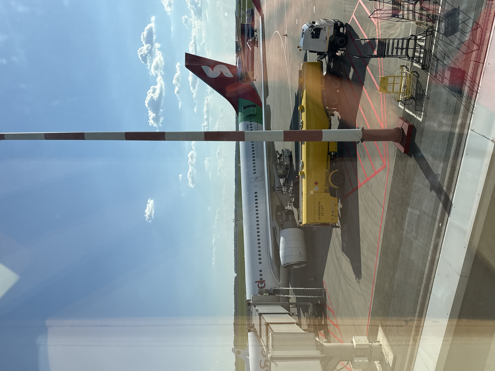 The image shows an airplane being refueled at what appears to be an airport gate. The aircraft has a red and green tail fin with a stylized "S" logo, likely identifying the airline as Nordwind Airlines. A yellow fuel truck is parked alongside the plane, with refueling equipment connected. Other airport service vehicles and equipment are visible in the background, including a possible de-icing apparatus. The environment is a typical airport tarmac, featuring painted lines on the concrete surface. The scene is captured from inside a building, possibly an airport terminal, with reflections visible on the glass. There is no indication that this picture was taken in Ufa, Russia.