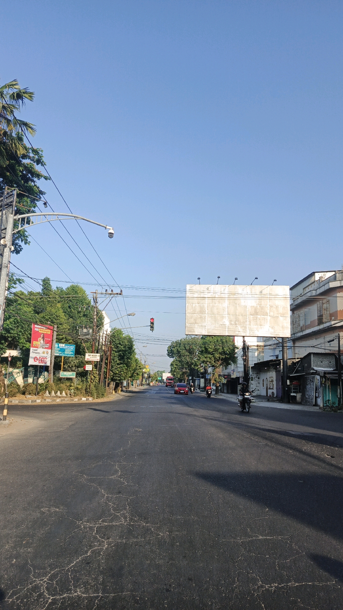 The image depicts a street scene in Surakarta, Indonesia, under a clear blue sky. The perspective is from a street level, looking down a moderately wide asphalt road. The road surface shows significant cracking and wear. On the left side of the road, there are trees, a utility pole with wires, a traffic camera, and several billboards and signs. One prominent red and white billboard on the left features text and a picture of a person. Further down the road on the left, there is a traffic light showing red.
On the right side of the road, there are buildings, including a multi-story structure with an orange and white facade. Several smaller structures with shopfronts line the street.
Vehicles are present on the road. Several motorcycles are visible, with riders on them. There are also a few cars, including a red car in the middle distance. The traffic appears to be light to moderate. The sun is casting long shadows across the road, indicating it is likely morning or late afternoon. The overall impression is a typical urban street in Indonesia during daylight hours.