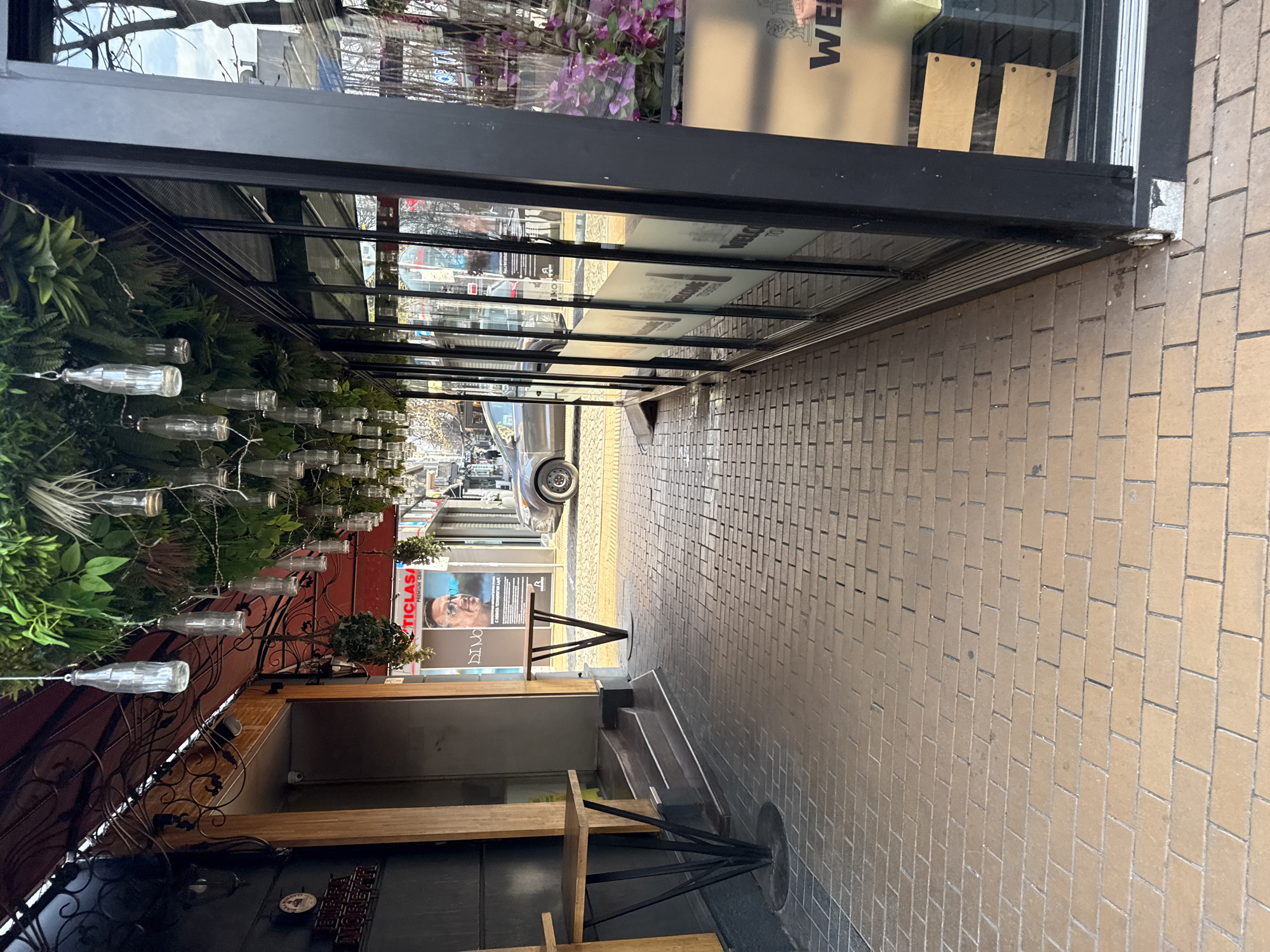 This image depicts a pedestrian walkway on Ulitsa Georgi S. Rakovski in Sofia, Bulgaria, identified by a visible "TICLASA" poster advertising a dental clinic known to be on this street. The perspective is longitudinal, looking down the path towards the street.
To the left, a building presents a dark facade with a red awning. Beneath the awning, black wrought iron decorative elements are affixed, alongside artificial green foliage and hanging planters. A "BIRRA MORETTI PUB" sign is partially visible on the wall. Two wooden-topped tables with black metal bases are positioned against this wall, flanking a short, two-step staircase leading to an elevated entrance. A security camera is mounted above this entrance.
Overhead, an artificial canopy of dense green foliage, including ferns, covers the walkway, from which numerous clear glass bottles, consistent in shape with vintage soda bottles, are suspended by thin wires at varying heights.
To the right, a modern commercial establishment features a floor-to-ceiling glass facade with black metal framing. The lower sections of the glass panes display frosted horizontal decals partially reading "WELCOME TO." Inside, artificial purple flowers are visible. Reflections in the glass show bare tree branches, distant buildings, and a hazy sky. The base of the glass structure shows minor external wear.
The walkway is paved with light-brown rectangular bricks in a running bond pattern, transitioning to a darker grey paving material near the left building. Beyond the immediate walkway, the street is paved with a lighter, textured material, possibly cobblestone. A grey sedan is parked on the street, partially visible and facing away. Further down the street, additional commercial storefronts and bare trees are observed. No individuals are clearly visible in the foreground or mid-ground. The scene is illuminated by diffuse daylight.