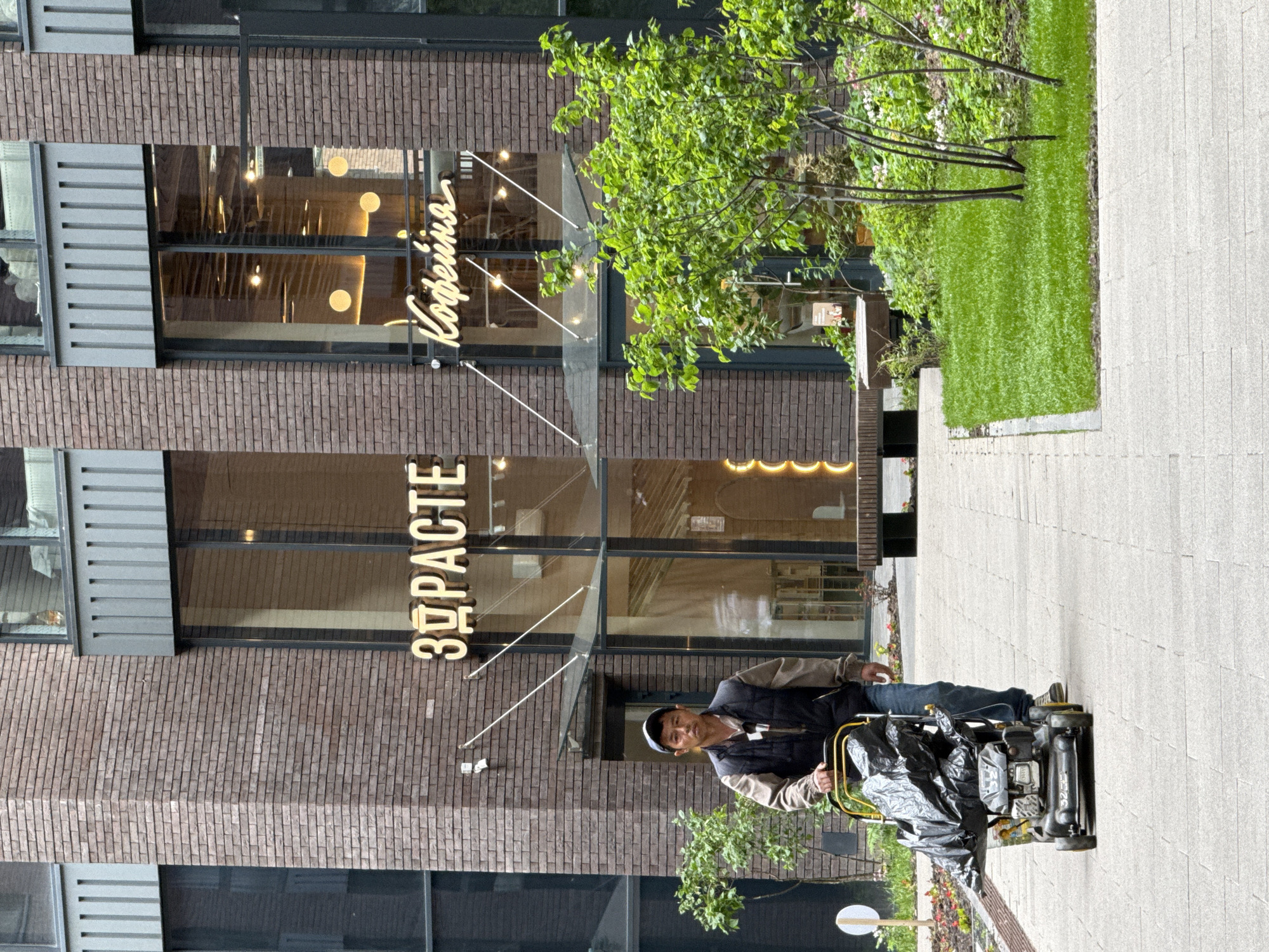This photograph captures an outdoor urban scene in Moscow, Russia, during daylight hours on what appears to be a clear day. The primary subject is a man standing on a light grey paved sidewalk, operating or pausing with a compact utility machine. He is dressed in a dark cap, a dark vest over a light long-sleeved shirt, and blue jeans, looking directly towards the camera with a neutral expression. The machine he is with has a yellow frame, black engine components, and a large black plastic bag attached, suggesting it's used for street cleaning or waste collection.
Behind the man is a modern building with a facade of dark reddish-brown bricks and large, clear glass windows on the ground floor. In front of the building, there is a small strip of green lawn and some young trees or bushes.
Two prominent illuminated signs are visible above the building's glass entrance. The sign on the left displays the Russian word "ЗДРАСТЕ" (ZDRAS'TE), an informal greeting meaning "hello." The sign on the right reads "Кофейня" (Kofeynya), which translates to "Coffee Shop." Both signs emit a warm, golden light. The visible text and modern urban architecture firmly place the scene in a contemporary Russian city setting, aligning with the provided context of Moscow.