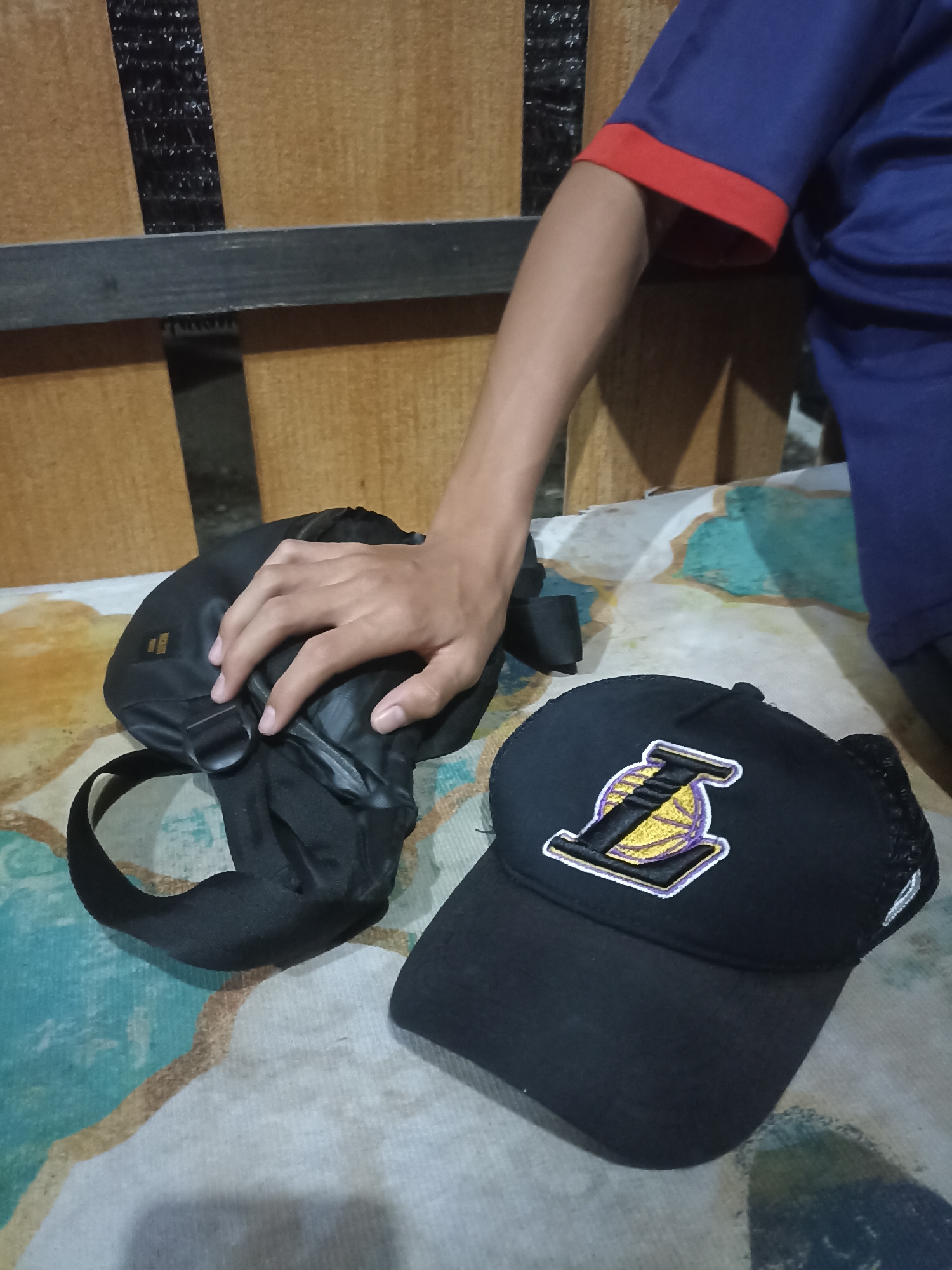 A person's arm and shoulder are visible on the right side of the image, wearing a blue t-shirt with red trim. A black fanny pack is placed on a patterned surface to the left of the person's hand, which is resting on it. Next to the fanny pack is a black baseball cap with a Los Angeles Lakers logo embroidered on the front. The background appears to be made of wooden planks with black tape. The scene is set in a room, likely within a home, in Mojopanggung, Indonesia.