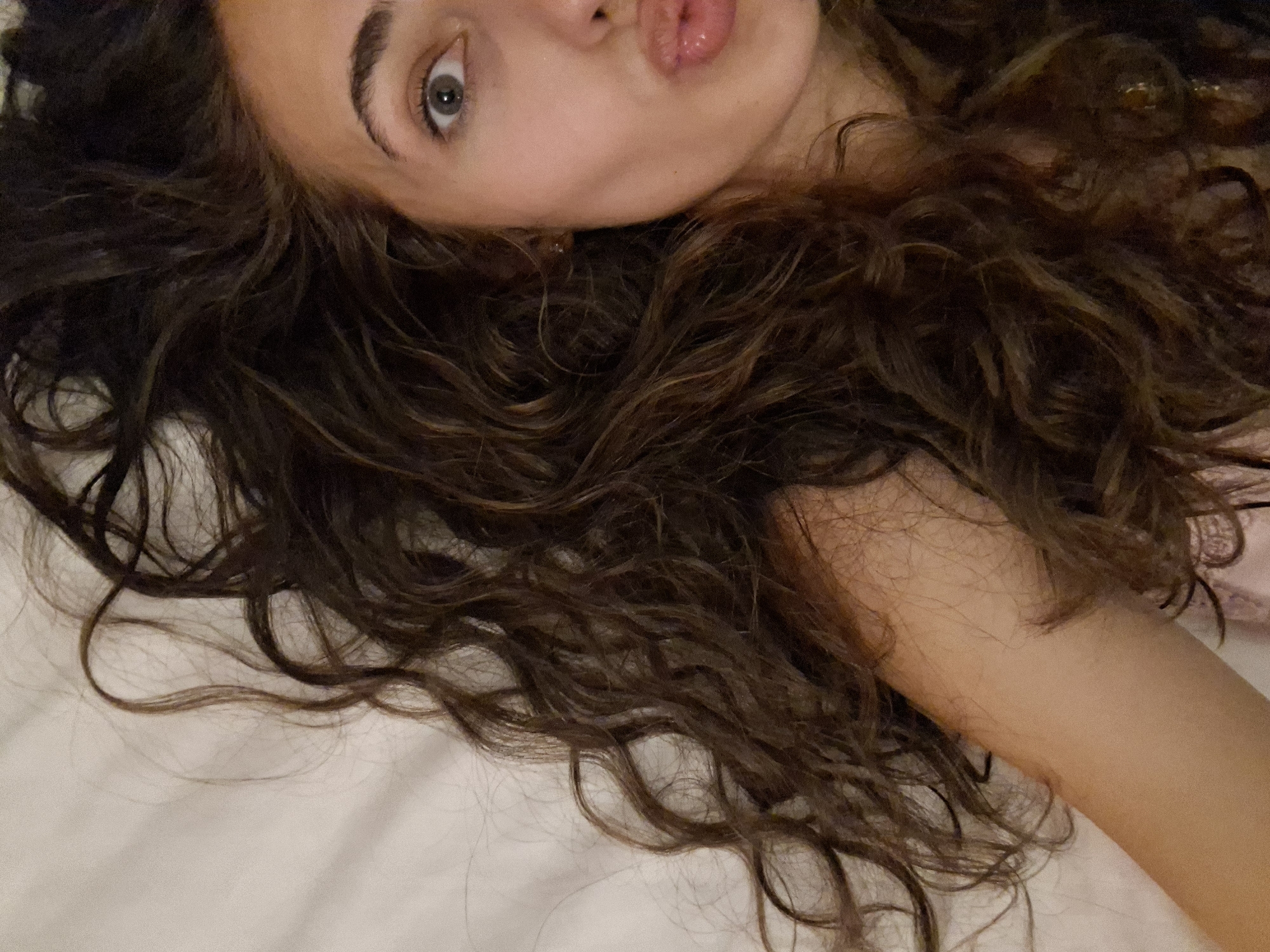 A close-up, overhead shot of a young woman with dark, curly hair lying on a white surface. Her head is angled towards the left side of the frame, with her right eye and lips visible. Her eye is open and looking towards the camera, and her lips are pursed as if for a kiss. Her hair is spread out around her head and shoulders, some strands falling onto the white surface. The background is a plain white surface, possibly a bed or a sheet. No other objects or people are visible in the frame. The lighting is soft and diffused, casting gentle shadows. The overall impression is one of a candid, relaxed moment.