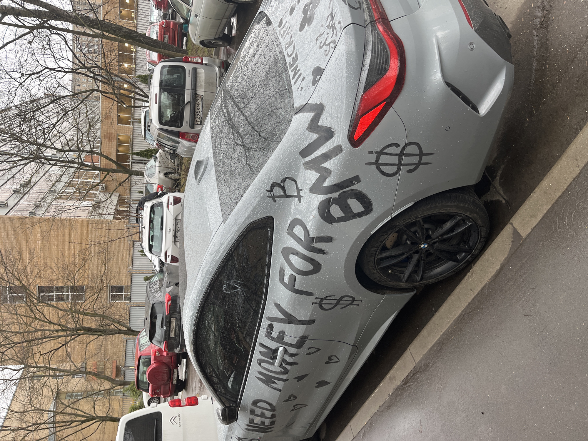 The image shows a light gray BMW with graffiti on its body. The graffiti includes the phrases "NEED MOHEY FOR BMW" and drawings of dollar signs, hearts, and the word "INECRAFTS". The car is parked on a street next to a curb. Other cars are parked in the background, including a white Citroen van and a red car. A multi-story brick building is visible in the background, along with bare trees. License plates indicate that the location is likely Moscow, Russia.