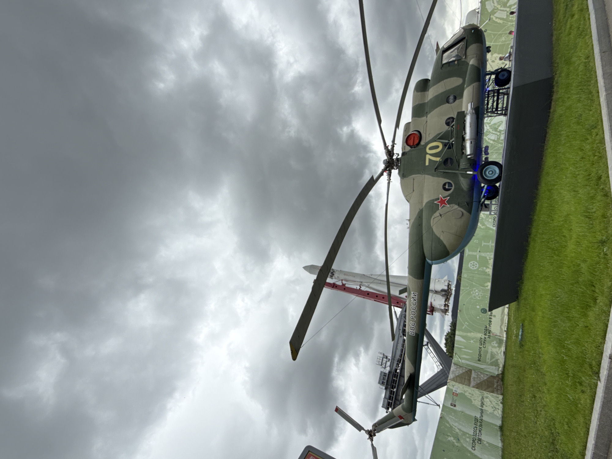 This media file features a vertical shot of a Russian military helicopter, likely a Mi-8 or Mi-17, on display. The helicopter is painted in a camouflage pattern of green and tan. The number "70" is prominently displayed on its side, along with a red star emblem. A red light is visible on the top of the helicopter's fuselage. In the background, a white rocket stands vertically, supported by a red crane. The scene is set outdoors under a cloudy, overcast sky, suggesting a cool or temperate day.
The foreground shows a patch of vibrant green grass and a dark, flat surface, possibly a display base or walkway. A light-colored barrier with printed text runs along the right side of the frame. The visible text, in Russian, appears to be promotional or informational for an upcoming attraction: "СКОРО ЗДЕСЬ БУДЕТ СВЕТОМУЗЫКАЛЬНЫЙ ФОНТАН" which translates to "SOON THERE WILL BE A LIGHT AND MUSIC FOUNTAIN HERE". Another piece of text reads "ВВС РОССИИ," meaning "AIR FORCE OF RUSSIA." Further text indicates "ВОДНОЕ ШОУ - СТРУИ ВОДЫ 35 МЕТРОВ В ВЫЛ," which translates to "WATER SHOW - WATER JETS 35 METERS IN HEIGHT." These inscriptions strongly suggest the location is an exhibition or park in Russia, likely Moscow, celebrating aviation and future attractions. The overall mood is solemn and informative, typical of a museum or exhibition setting.