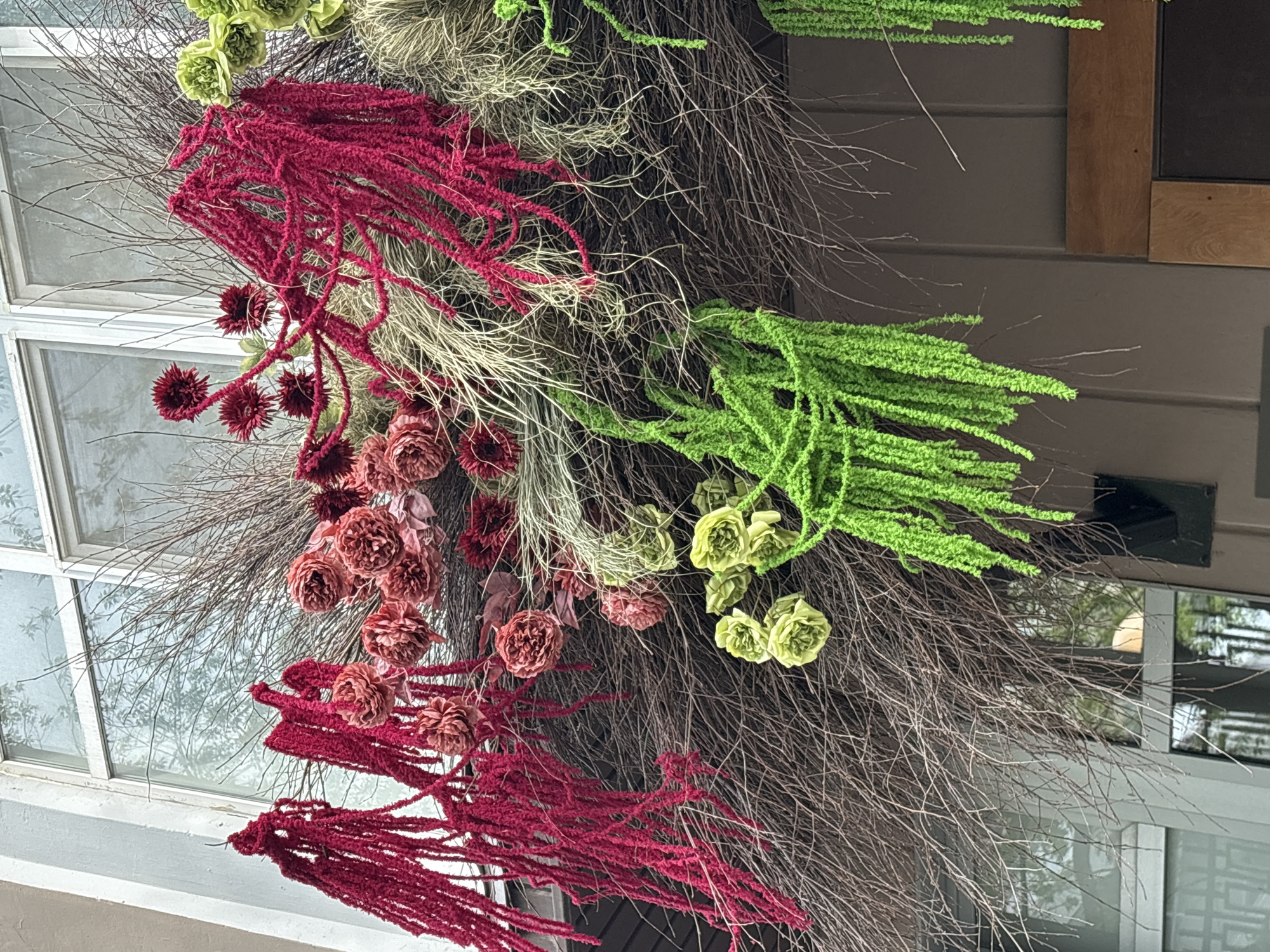 A close-up shot shows a decorative floral arrangement hanging on a wall near a window. The arrangement features red and green amaranthus, brown dried branches, and beige Spanish moss. The window and the wall it is mounted to are not in focus, but they give a sense of location with the architectural style. The location is Perm, Russia.