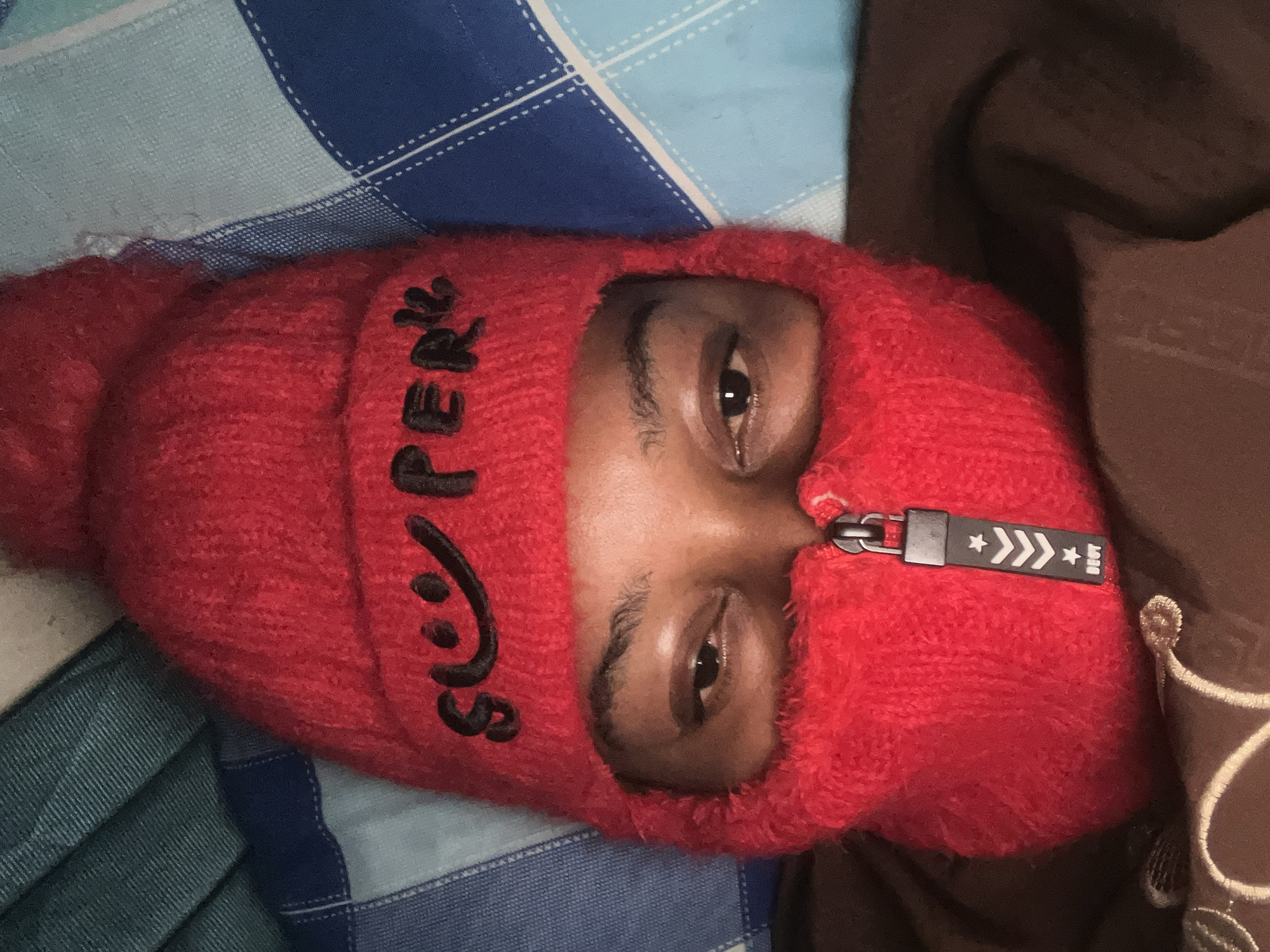 A close-up shot shows a person lying down, wearing a red knitted balaclava with the word "SUPER" embroidered in black on the forehead. The balaclava has a zipper in the front that is partially open, revealing the person's eyes and eyebrows. The background consists of a blue and white checkered fabric, likely a bedsheet. The lighting suggests an indoor setting. No specific location in Jos, Nigeria, can be identified from the image.