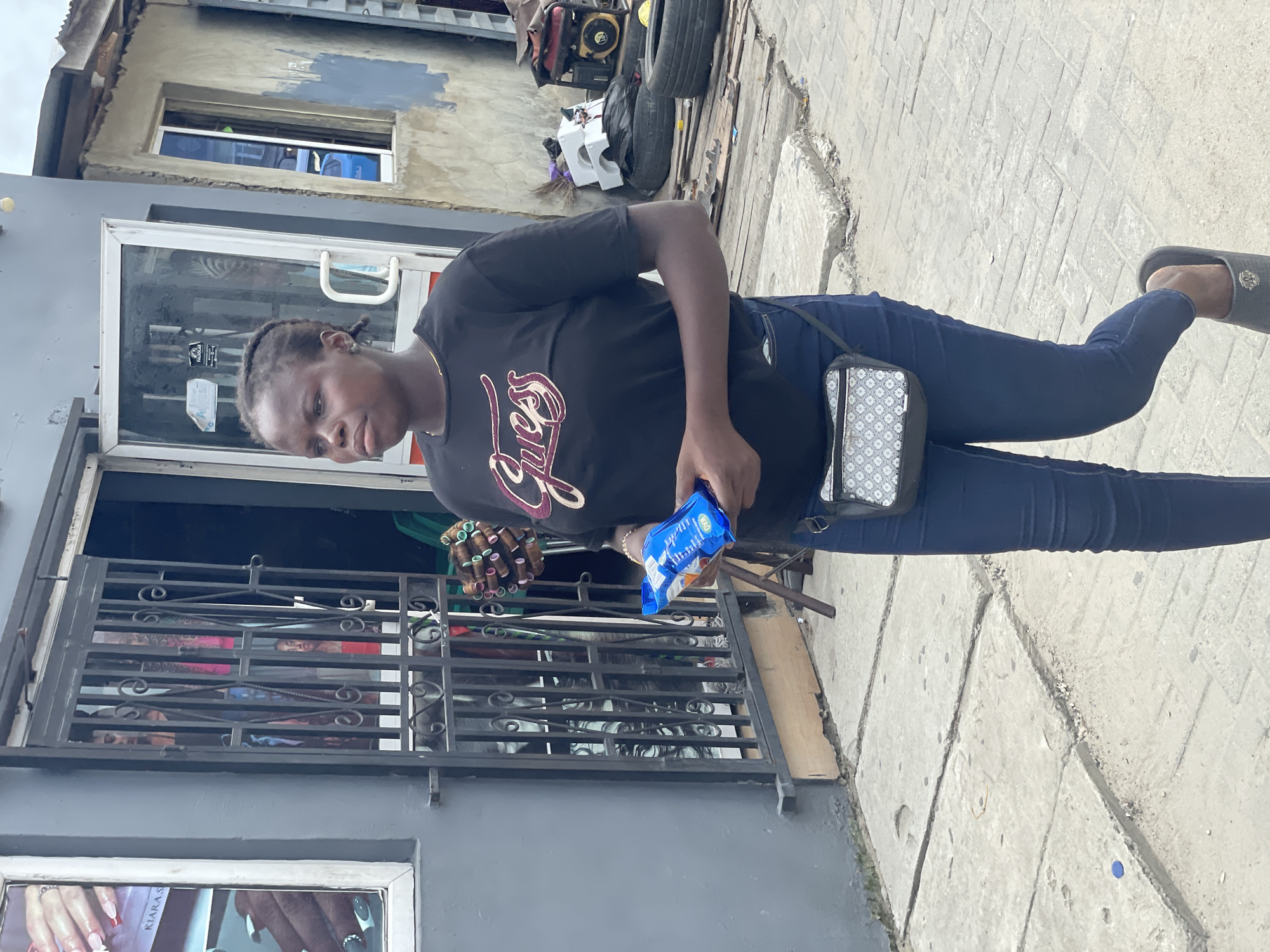 A woman stands on a concrete sidewalk near a building in an urban location, likely a city in an unknown country. The woman wears a black t-shirt with the word "Guess" written on it, blue jeans, and dark sandals. She has a small crossbody bag and is holding a blue snack package. The building is painted a light grey color and features a storefront with a metal security gate and a glass door. Other items such as a generator, tires, and miscellaneous objects are visible in the background. The sky appears overcast.