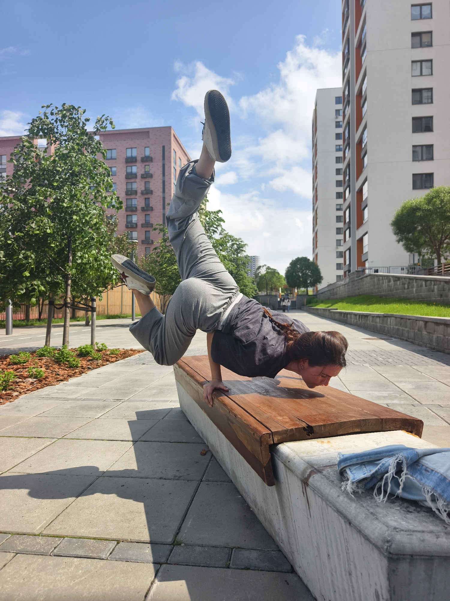 A woman is performing a handstand pose on a wooden bench in a city environment. She is wearing gray pants, a dark gray t-shirt, and sneakers. Her legs are extended upwards, forming a near-vertical line. The bench rests on a concrete base, with a blue denim jacket tossed to the side. The location appears to be a public space in Tyumen, Russia, characterized by paved walkways lined with trees and mulch beds. In the background are multi-story residential buildings, one with a pink brick facade and another with white and orange accents. The sky is blue with scattered clouds, indicating a sunny day.