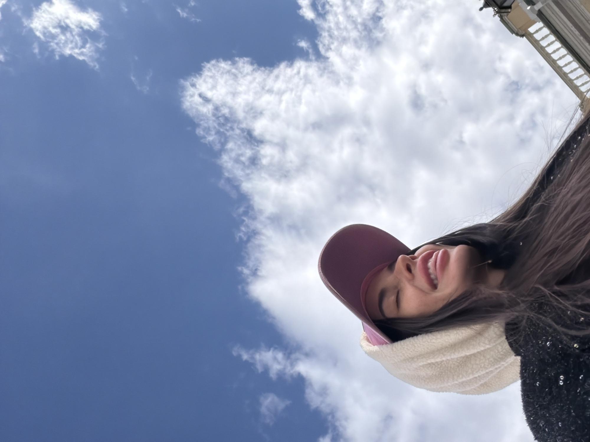 The image, captured from a low-angle perspective, features a young woman positioned in the lower-right quadrant, playfully looking towards the upper-left. She has long dark hair and wears a pink baseball cap and a cream-colored, fluffy neck warmer, with a dark, possibly sequined, garment visible on her right shoulder. Her eyes are closed, and her mouth is open with her tongue slightly extended, conveying a cheerful and lighthearted emotion, indicative of a fun moment or a selfie.
The background is dominated by a bright blue sky with scattered white clouds, primarily filling the upper-left and central portions of the frame. In the upper-right corner, a section of a light-colored architectural structure with visible columns or balustrades can be seen. This architectural detail, combined with the given location context, suggests a significant building or monument in Saint Petersburg, Russia. The scene is set outdoors during the daytime under a partly cloudy sky. No text is visible in the image.