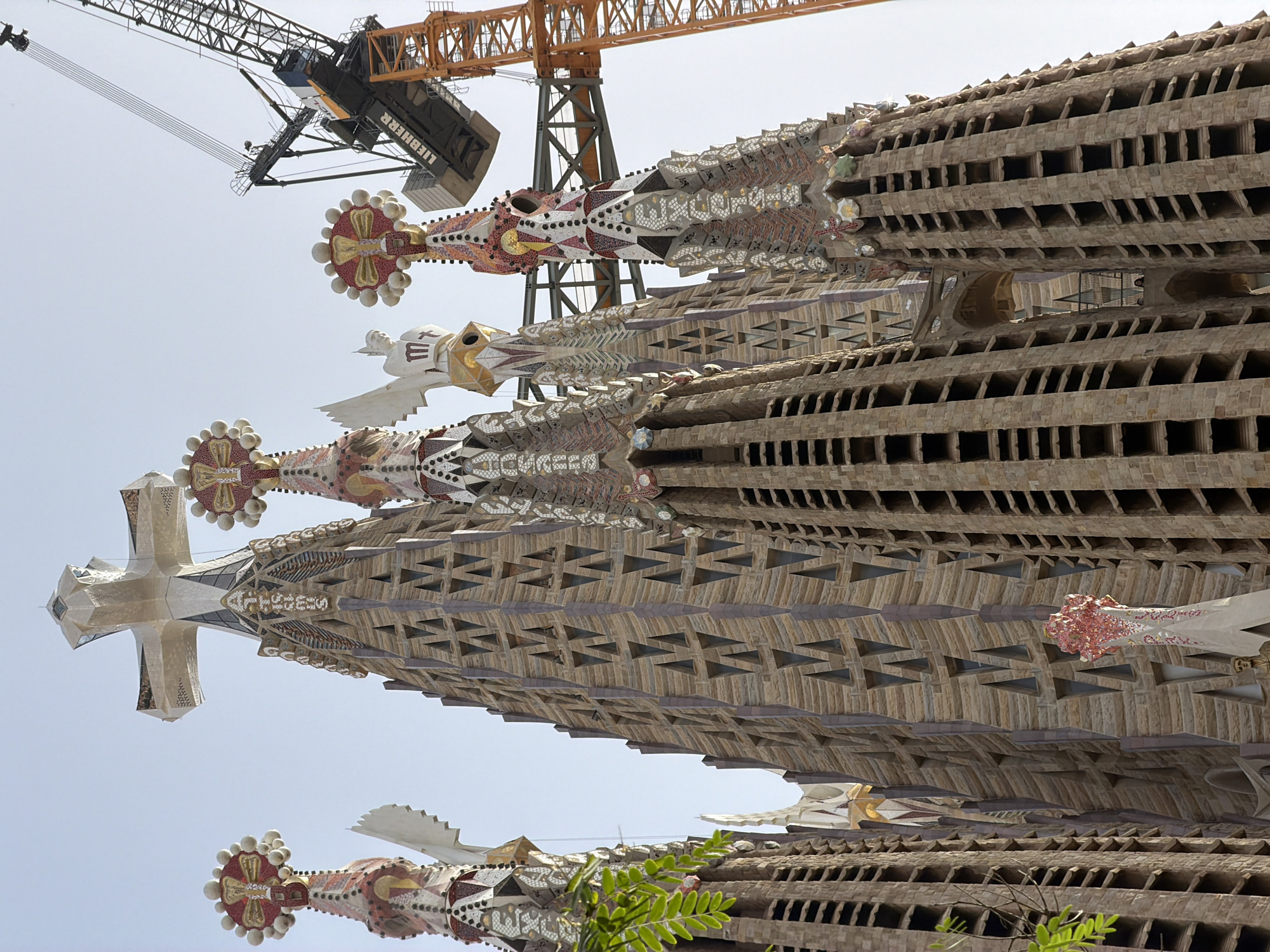 The image shows an exterior, low-angle, wide shot of the Sagrada Familia basilica in Barcelona, Spain. The sky is clear and bright. The basilica's distinctive spires, adorned with mosaic and sculptural details, are prominently featured. In the background, a large construction crane with the brand "LIEBHERR" visible on its arm is actively working on the basilica. Several towers are visible in various stages of completion, some with scaffolding and construction materials. The detailed stone architecture and the vibrant decorative elements of the spires are evident, highlighting the ongoing construction and grandeur of the building.