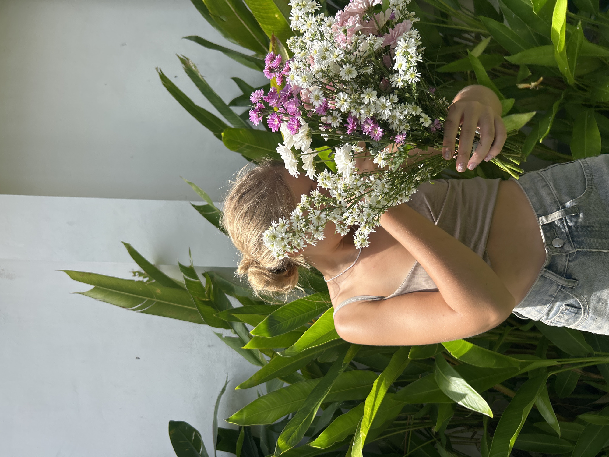 The media file displays a bright, outdoor scene featuring a person depicted horizontally across the frame. The subject, a woman with blonde hair styled in a bun, has her face largely obscured by a large, vibrant bouquet of flowers. She is wearing a light beige or cream-colored tank top and light blue denim shorts or jeans, with her upper torso and legs visible.
The prominent bouquet consists of numerous small, delicate white and purple-pink blossoms, resembling aster or similar daisy-like flowers, with green foliage intertwined. Her hands, adorned with light-colored nail polish, grasp the stems of the bouquet.
Behind the person, lush green tropical foliage with large, elongated leaves fills the lower and right portions of the frame. A plain white or off-white wall forms the background on the upper and left sides. The scene is illuminated by bright natural sunlight, creating clear shadows and highlights, indicative of a sunny daytime environment. There is no visible text in the image. The overall impression conveyed is one of natural beauty and tranquility, set in what appears to be a garden or outdoor space.