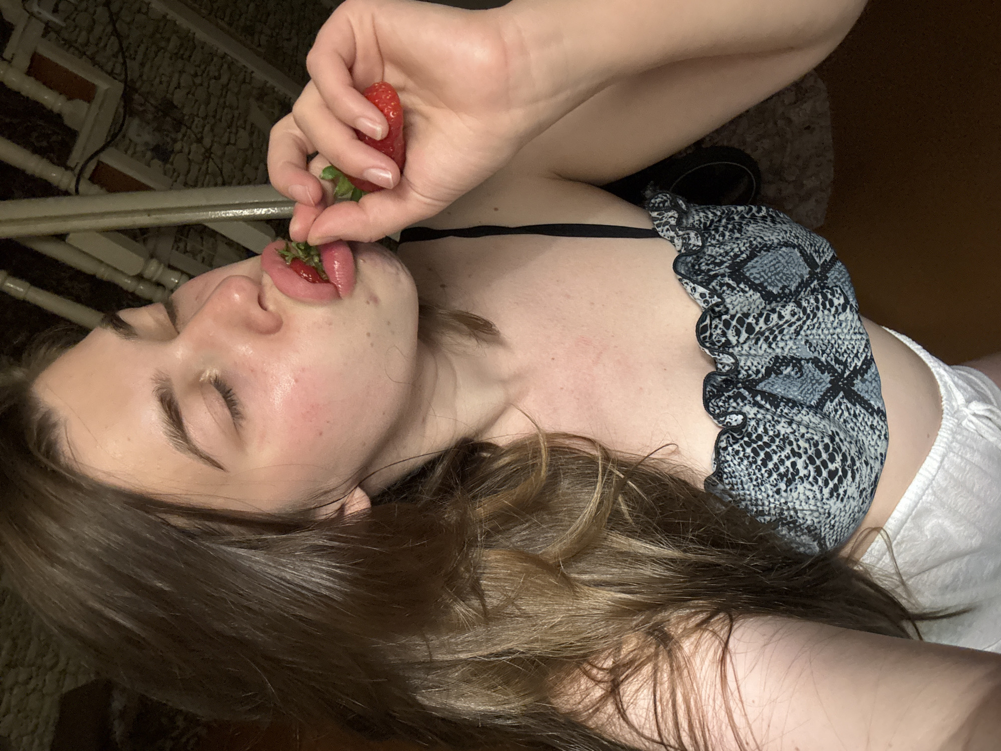 A woman with long brown hair is holding a strawberry to her lips. She is lying on her back and wearing a grey and black snakeskin-print top. Her eyes are closed and she is touching the strawberry to her tongue. The location is unknown.