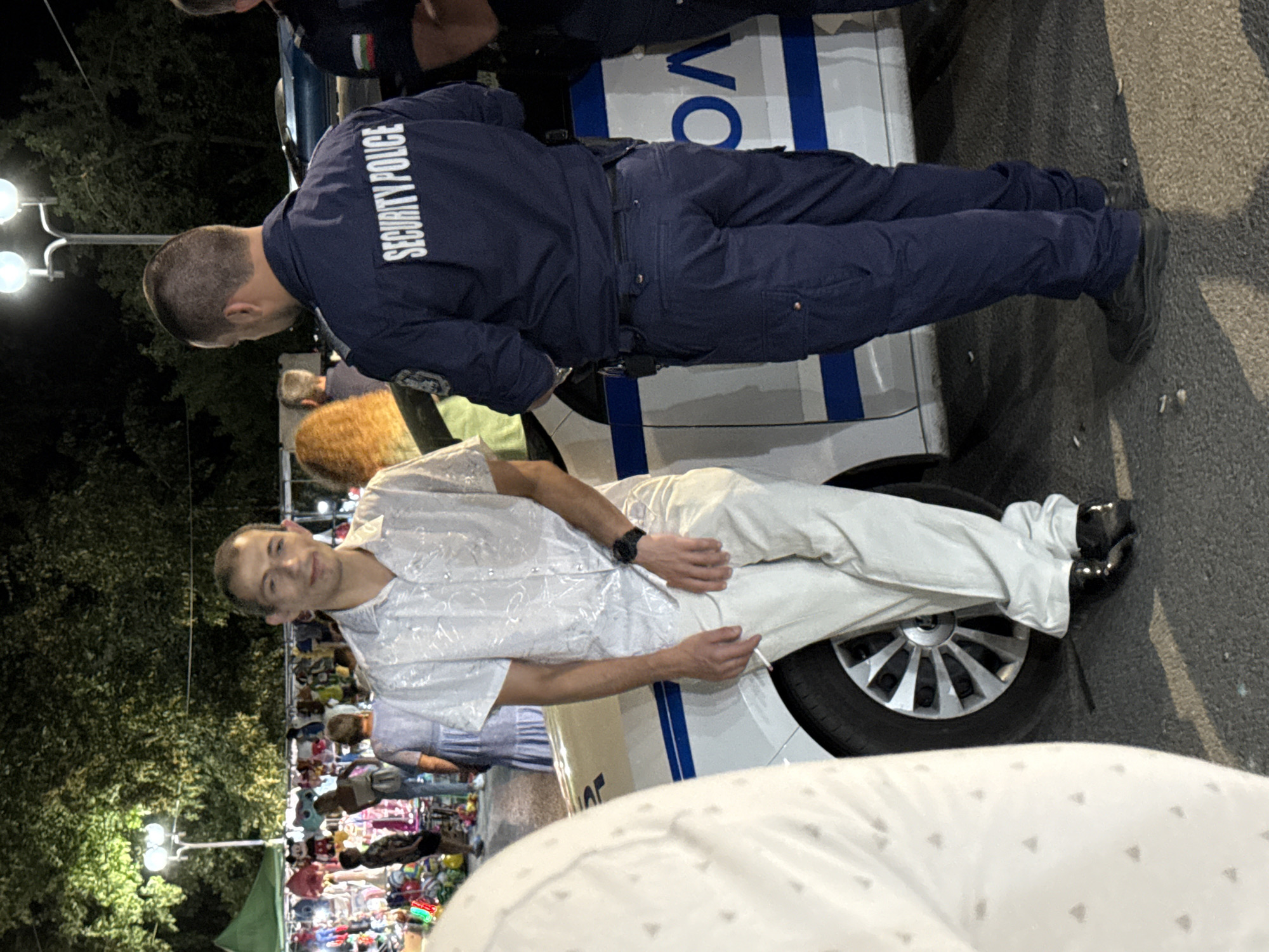 An outdoor scene at night, possibly a market or festival, in Stob, Bulgaria. A man in a white, patterned short-sleeved shirt and light-colored pants leans against a white car with blue stripes and the word "POLICE" visible. He is holding a cigarette. He wears a dark watch. Standing next to him, facing away from the camera, is a person in a navy blue uniform with "SECURITY POLICE" written on the back. The uniform includes a badge on the left sleeve and dark pants. The background shows trees, lighting fixtures, and stalls with merchandise, along with other people milling about.