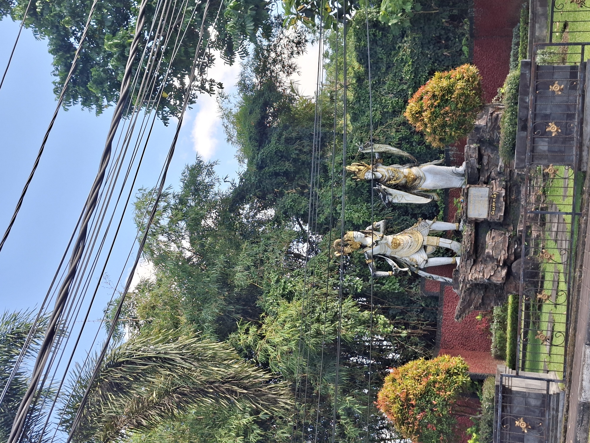 Two statues of what appear to be deities stand on a rock formation in front of dense green foliage. The statue on the left is in a dynamic pose, holding a sword and shield. The statue on the right is also adorned in regal attire. Both statues are white with gold accents. In the foreground, a black metal fence with gold decorative elements encloses a grassy area. Two small trees with red and green foliage are planted on either side of the statues. A red textured wall forms the background behind the statues and foliage. Power lines are strung across the upper portion of the image, against a clear blue sky with scattered clouds. The location is identified as Banjarangkan, Indonesia, with a sign at the base of the rock formation reading "DUMARA."