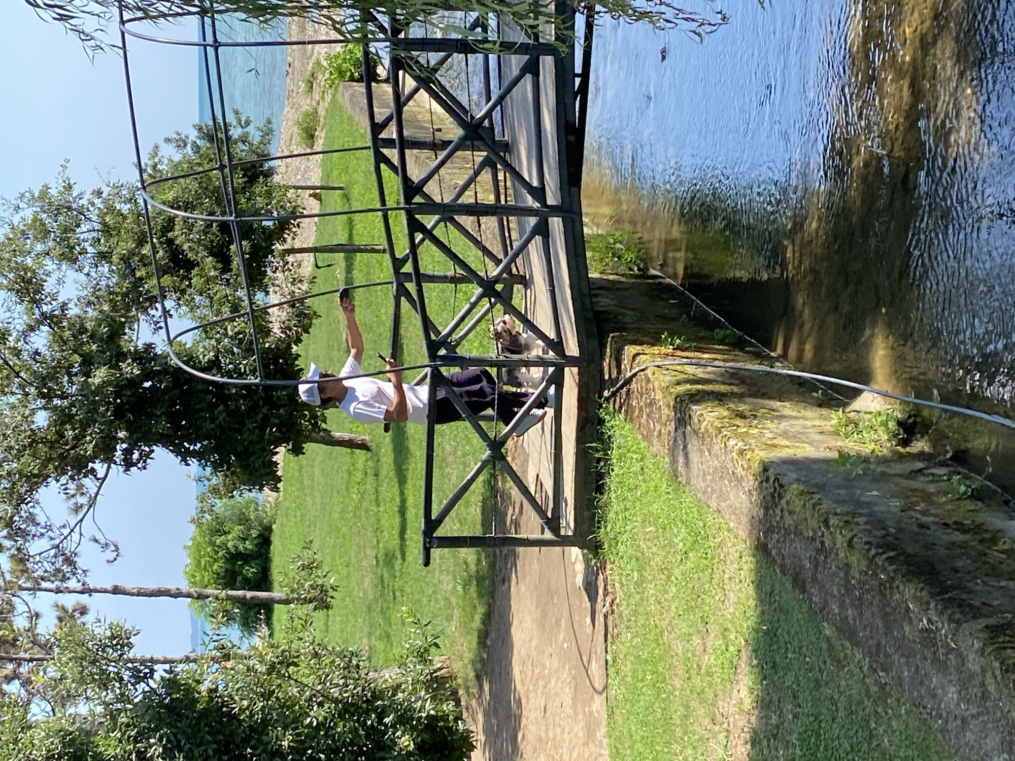 The image depicts a person holding a leash and walking a small dog across a wooden bridge spanning a narrow waterway. The person, wearing a white t-shirt and a light-colored cap, is looking down at the dog, which is a terrier mix with brown and white fur. The bridge has dark metal railings with a decorative criss-cross pattern. The waterway has clear, flowing water with some debris visible.
The surrounding environment is lush and green, with well-maintained grass and trees. In the background, beyond a line of trees, a body of water, likely the sea, is visible under a clear blue sky. The stone embankment lining the waterway is covered in moss and some small plants. This scene is consistent with the aesthetic of the Botanical Garden in Sokhumi, Abkhazia, known for its picturesque landscapes and diverse flora. The presence of the sea in the background further supports this location.