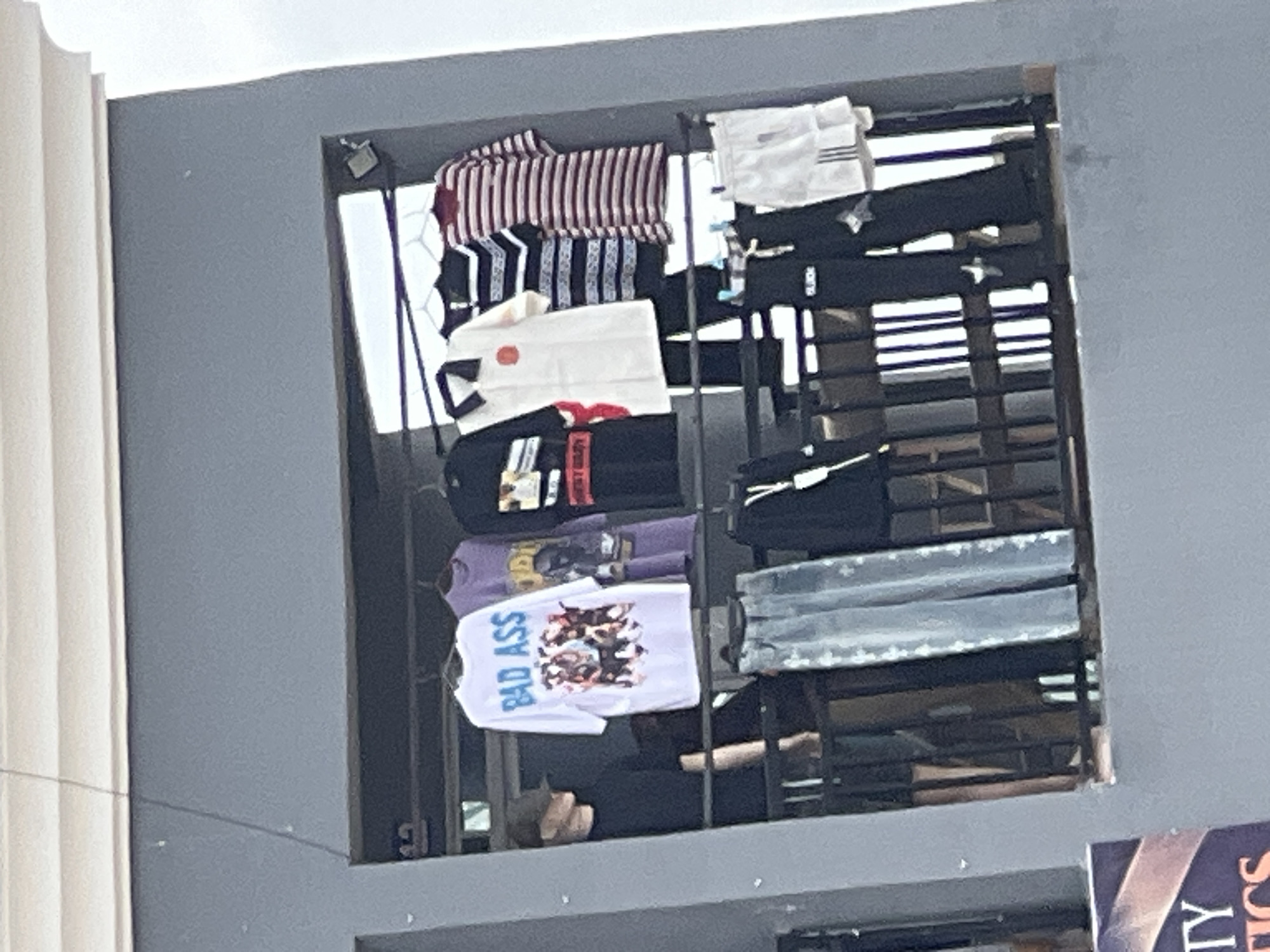 A person wearing a dark shirt and a cap stands to the left in front of clothing displayed on a balcony. On the balcony, several t-shirts and other apparel are hanging. From left to right, the shirts include a white t-shirt with blue "BAD ASS" text and a graphic, a purple t-shirt with a graphic, a black t-shirt with a graphic, a white polo shirt with a red emblem, a black and white striped long-sleeved shirt, and a red and white striped t-shirt. Below these, a pair of light-colored denim jeans is hanging, as well as dark pants and white shorts with black stripes. The balcony has a dark metal railing. The building appears to be in the city of Jalingo, Nigeria, as indicated by the presence of a sign in the lower left corner that reads "TY TICS" which likely refers to a business in Jalingo.