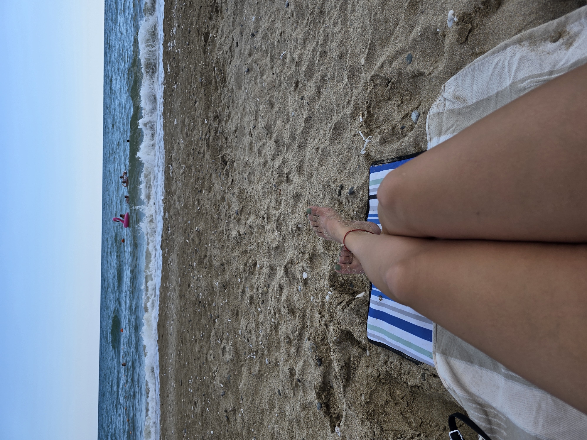 In Yevpatoriya, Ukraine, a person's legs are visible from the knees down, resting on a striped beach towel on a sandy beach. The person is lying down, facing the sea. Their feet are bare, with green nail polish on the toenails, and a thin red string is tied around one ankle. In the background, the sea is visible with gentle waves breaking on the shore. Several people are in the water, some swimming and others on inflatable toys, including a pink flamingo. The sky is clear and blue. The sand on the beach is speckled with small shells and pebbles.