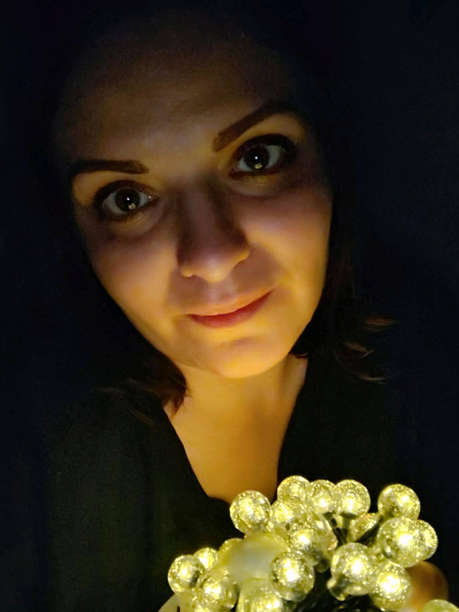The image is a close-up, low-angle, self-portrait of a woman holding a string of yellow fairy lights. She is looking directly at the camera with a slight smile. The lighting is dim, with the fairy lights illuminating her face and creating a warm glow. The background is dark and out of focus. There are no visible landmarks that would indicate a specific location in Марковский, Russia. The woman appears to be in an indoor setting.
