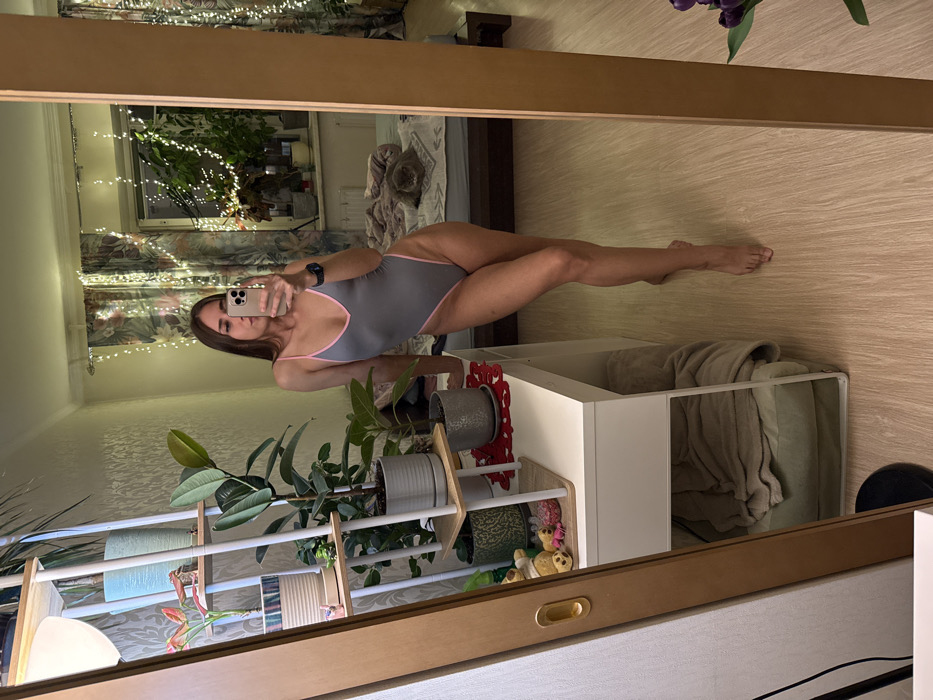 This image captures an interior room, identified as a residence in Saint Petersburg, Russia, through a full-length mirror selfie. A female, appearing to be in her 20s or 30s, stands barefoot. She is dressed in a grey one-piece swimsuit with pink trim, holding a light-colored smartphone (likely an iPhone) in her right hand, extended to take the photograph. Her left forearm rests on a white desk situated in the foreground. A smartwatch is visible on her left wrist.
The reflected environment includes a white multi-tiered shelving unit to the left, displaying multiple potted plants in various containers. On the white desk, a red patterned mat is visible. Beneath the desk, a green pet bed with a beige blanket is situated. In the background, a bed with white and blue bedding features a grey cat resting on it. A window behind the bed is adorned with floral curtains and multiple strands of white string lights. Additional potted plants are present on the windowsill. The room's floor is light-toned wooden or laminate, and the walls have a textured wallpaper.