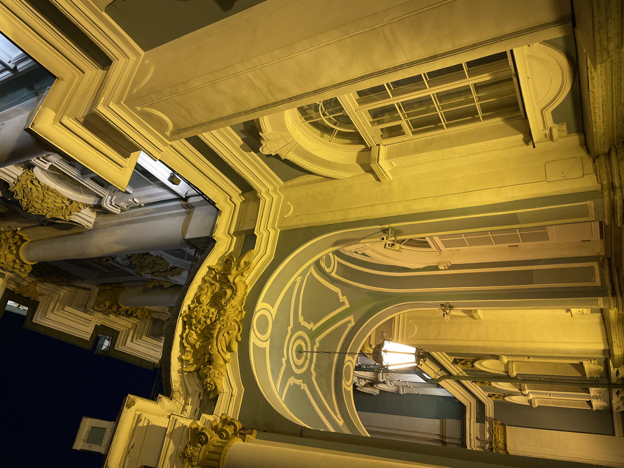 This media file captures an upward view of an ornate architectural structure at night, showcasing intricate details under warm artificial lighting. The scene is devoid of people and activity, focusing solely on the building's aesthetic.
The perspective looks up into a complex arrangement of arches, moldings, and decorated surfaces. The dominant elements are illuminated in a golden-yellow hue, highlighting their textures and forms. Intricate carvings, possibly in a Baroque or Neoclassical style, adorn cornices and pilasters, featuring organic motifs such as floral patterns or scrolling foliage. Sections of the structure are painted in a muted green or teal color, contrasted by cream or white stone elements.
Several windows with grid patterns are visible, some arched and others featuring circular insets. A hanging, illuminated lantern with a classic design is suspended from one of the archways. The dark, unlit sky is visible in the background, reinforcing the nighttime setting. This architectural display suggests a grand public building or a historical palace, consistent with the provided location context of Saint Petersburg, Russia. No discernible text is visible within the image.