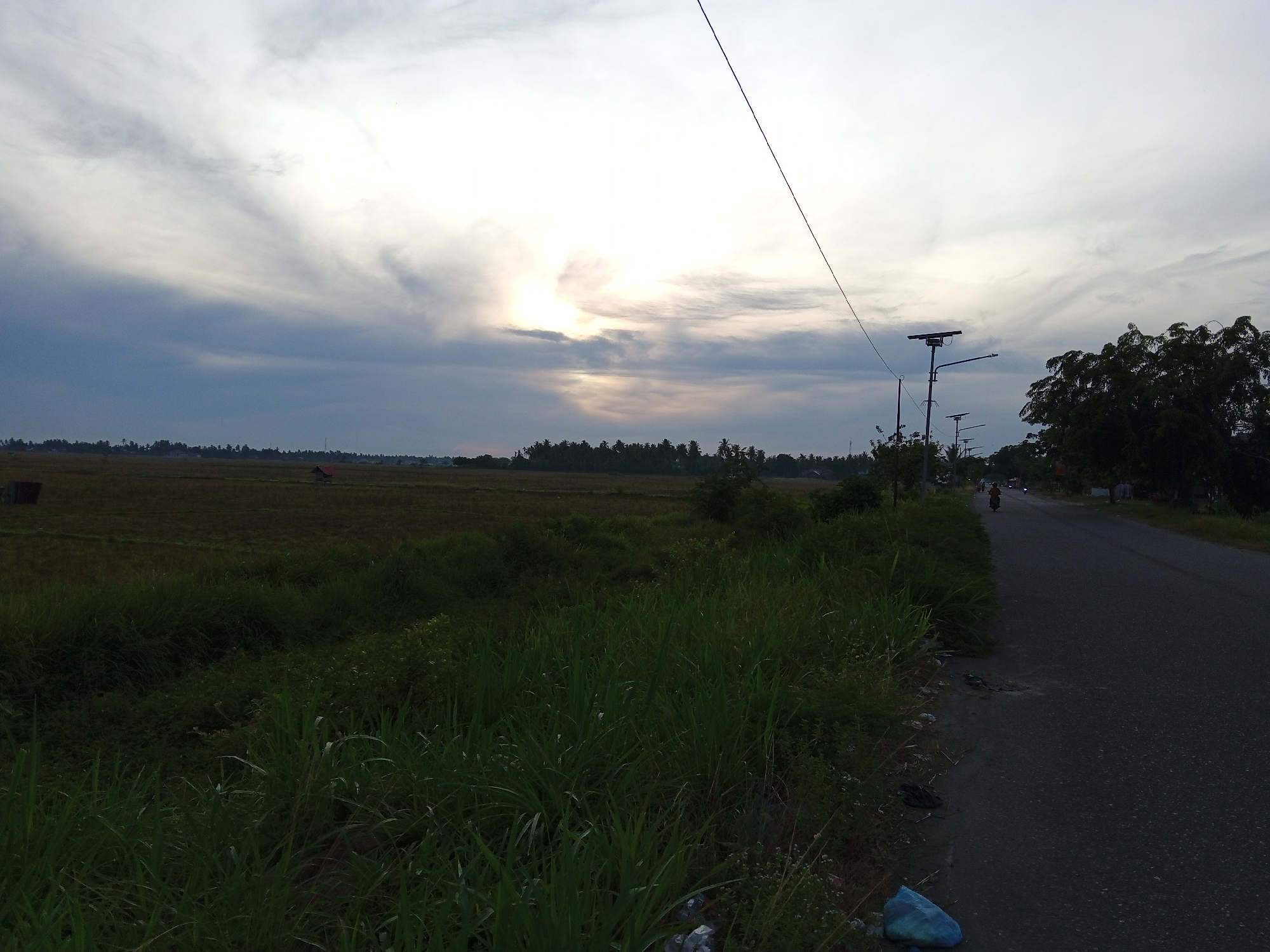 The image depicts a rural roadside scene in Cotgapu, Indonesia, likely during late afternoon or early evening, judging by the sky conditions and lighting. The sky is overcast with dramatic cloud formations and a break where sunlight is filtering through, creating a bright spot. A single utility pole with a solar panel is visible along the right side of the road, and several other similar poles recede into the distance.
To the left of the road is a field of tall, dark green grass and vegetation, with a hint of a cultivated field or rice paddy further in the background. A small red-roofed structure is visible in the distance within the field, possibly a small building or shelter. A dark asphalt road stretches into the distance on the right, with a single motorcycle with a rider heading away from the viewer. The edge of the road is littered with some plastic bags and debris. A dense cluster of trees with lush green foliage borders the right side of the road further ahead. The overall impression is one of a quiet, undeveloped area on the outskirts of a settlement.